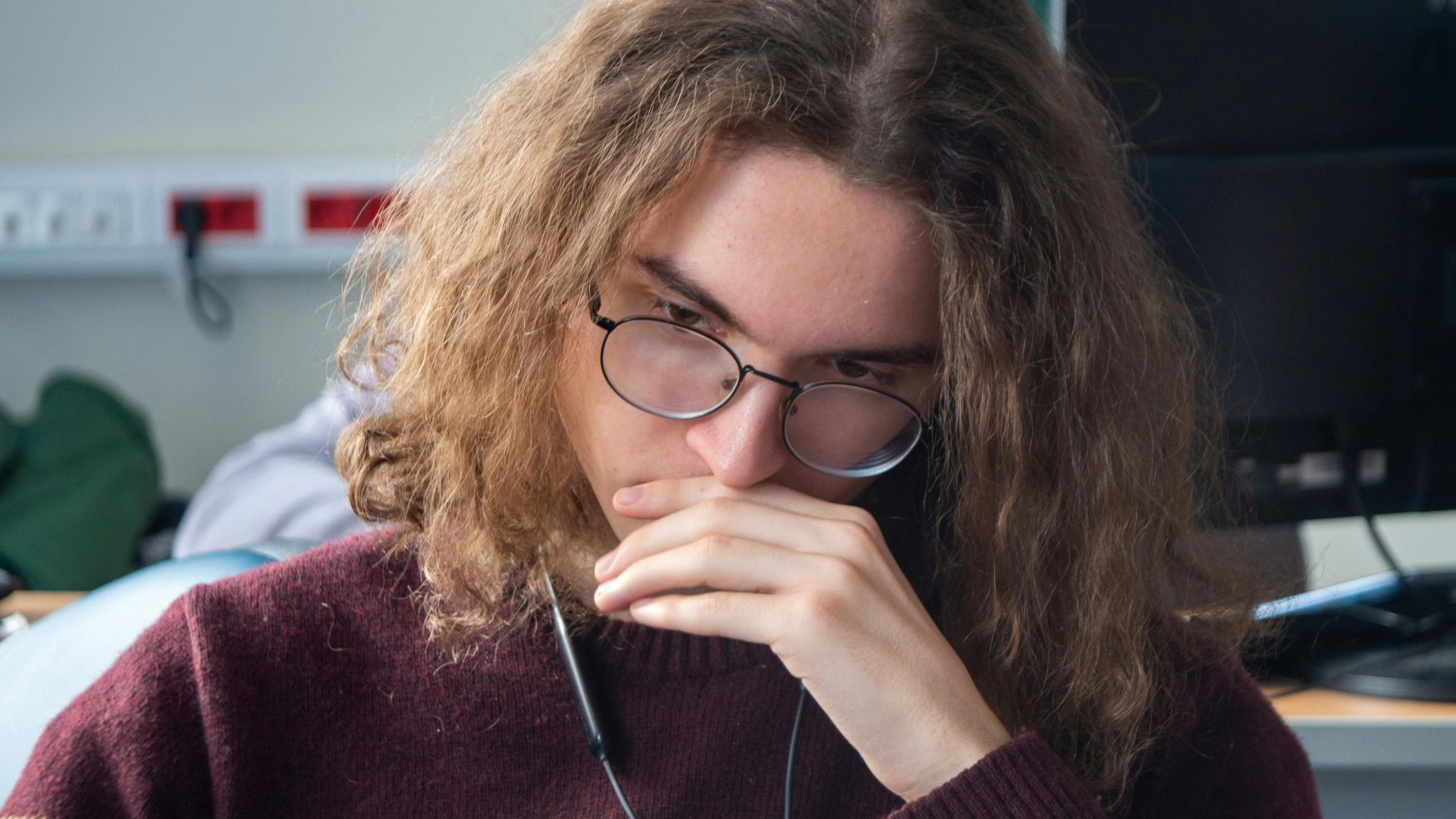 Young adult wearing glasses and sweater, deep in thought at a workstation.