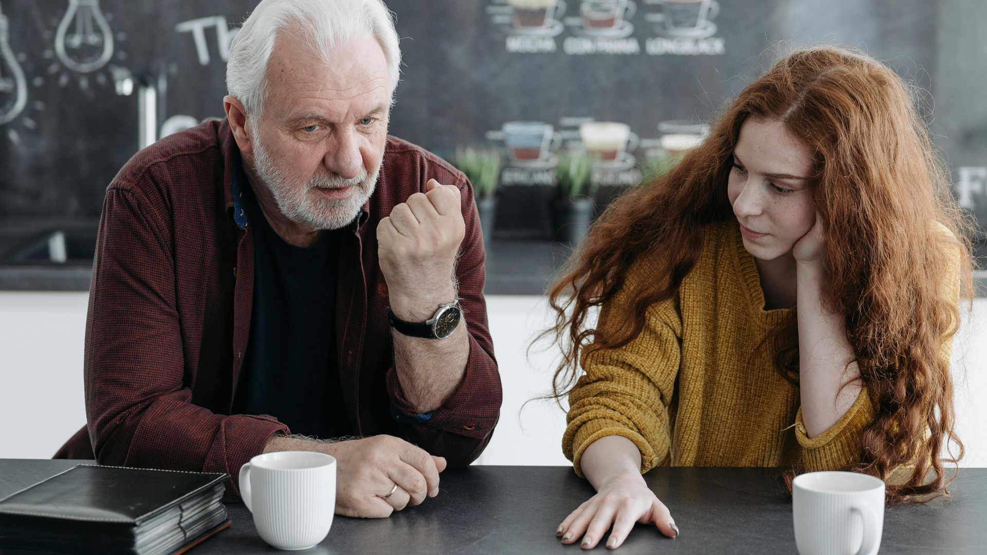 An elderly man and his granddaughter engage in a heartfelt conversation over coffee indoors.
