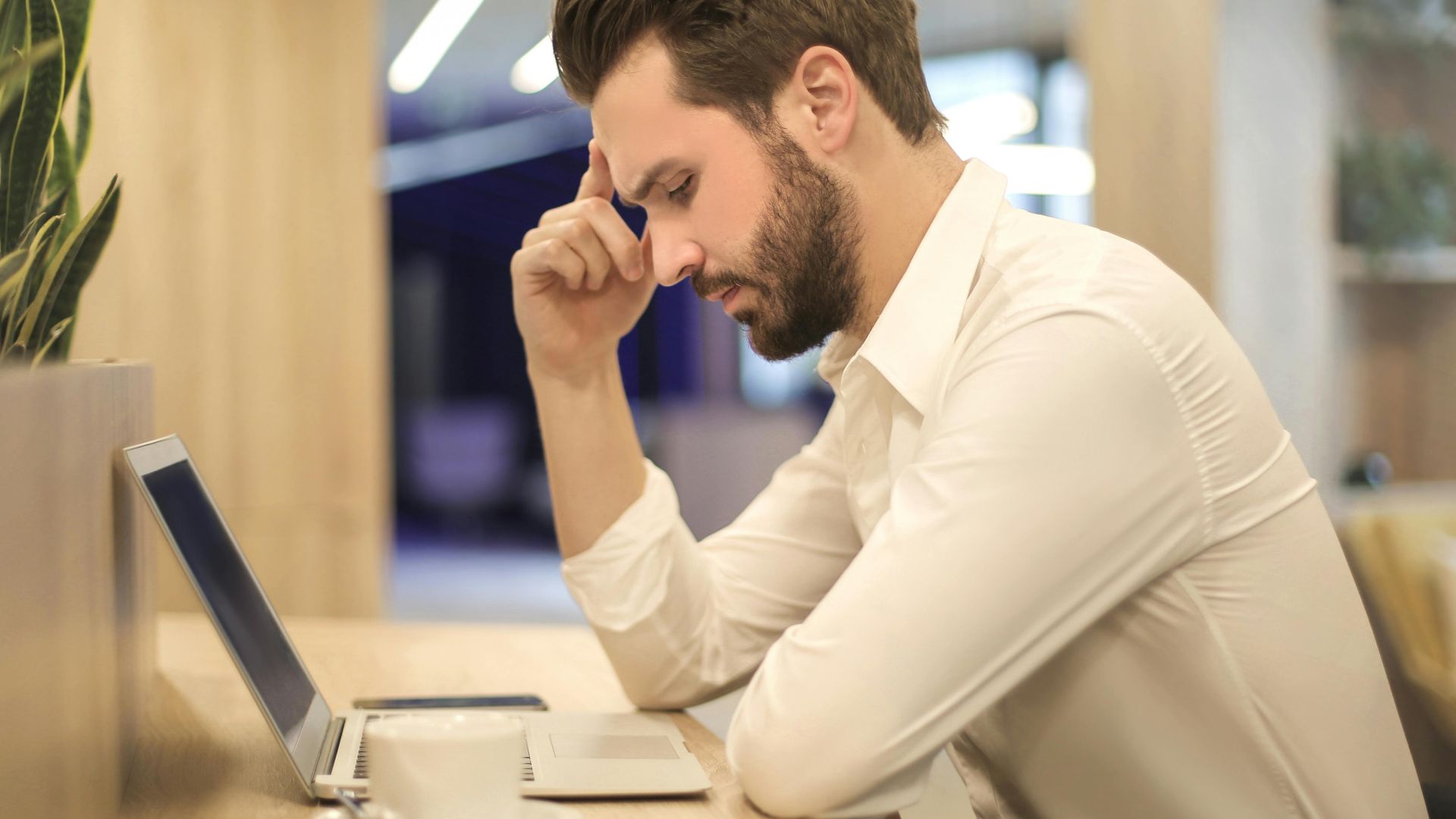 A thoughtful man in a shirt works on his laptop at a modern indoor office space.