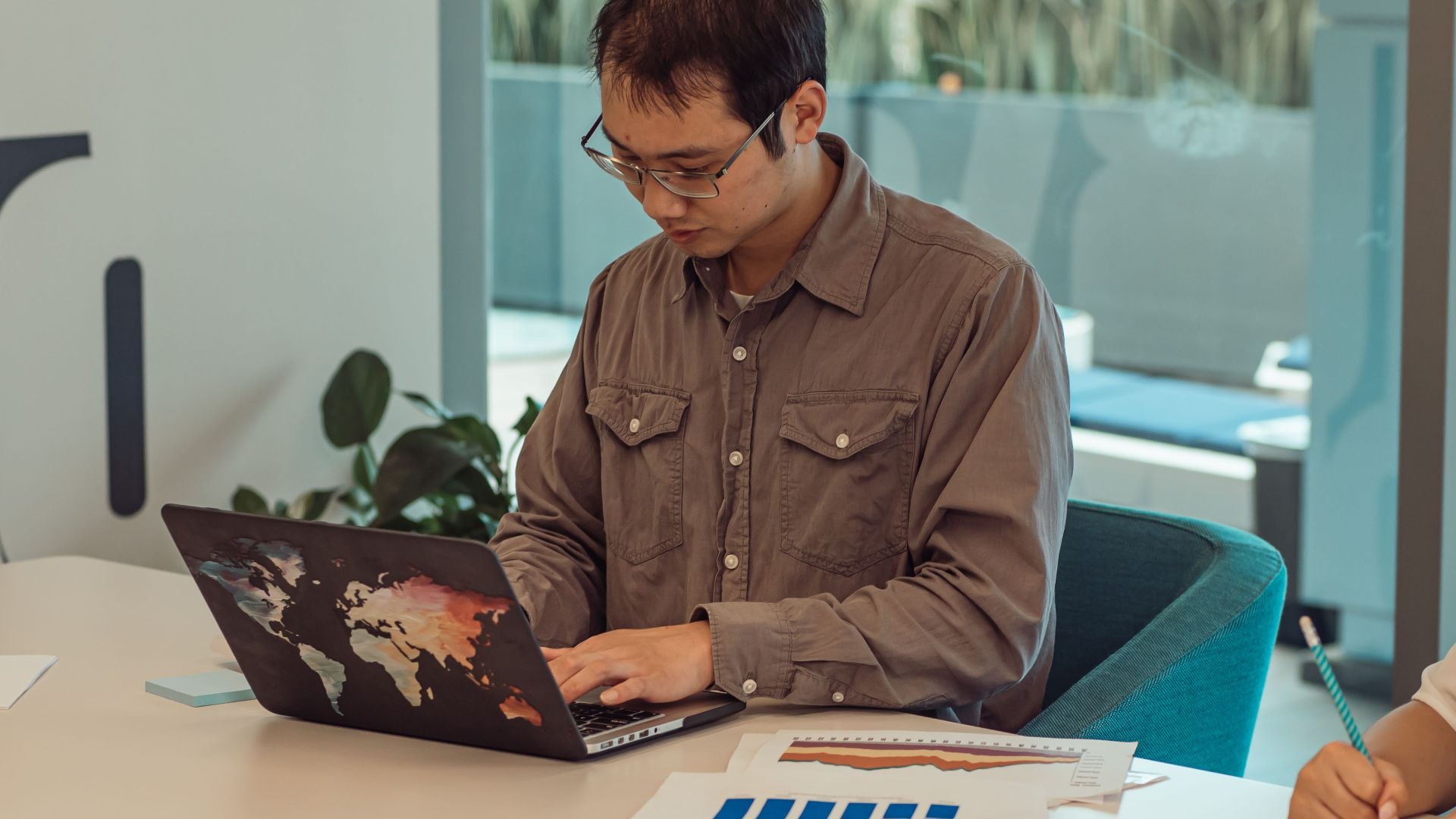 Focused Asian man working on a laptop surrounded by papers in a modern office setting.