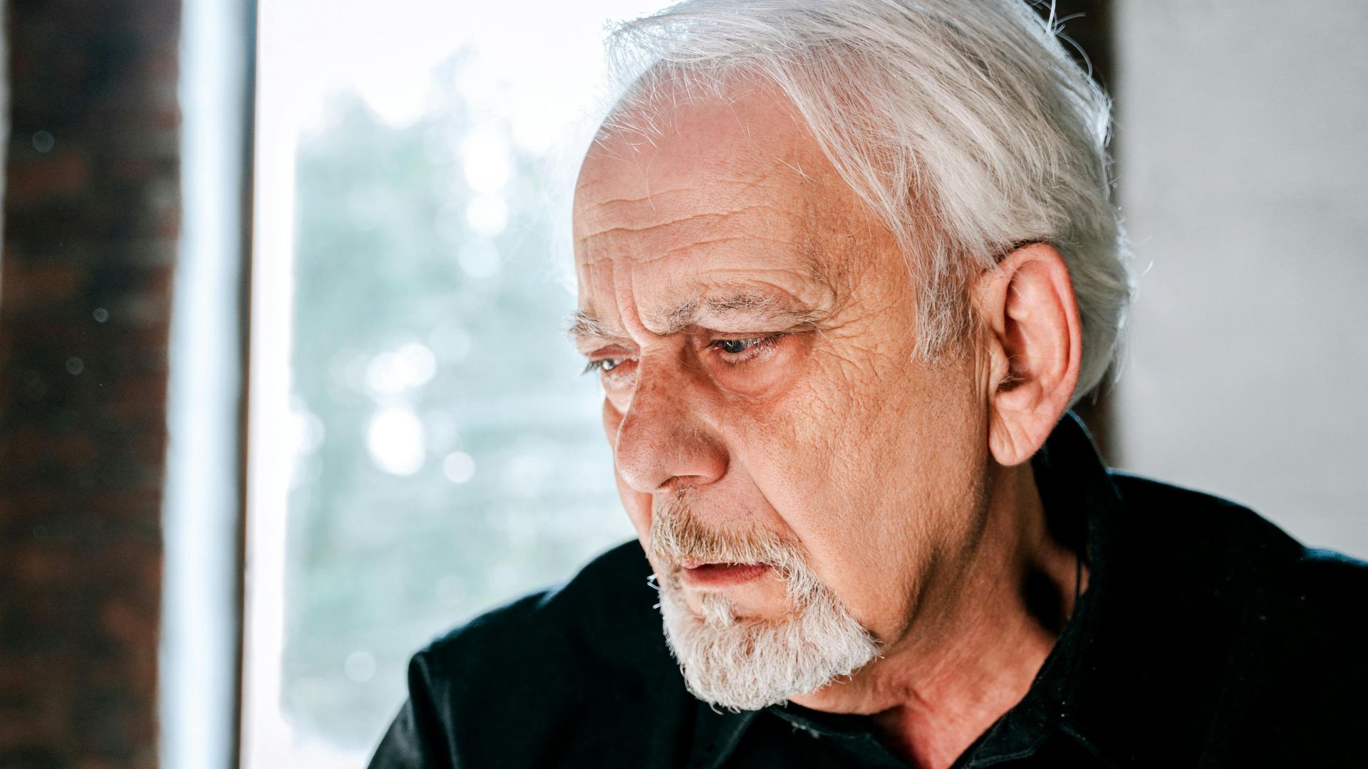 Pensive elderly man with white beard in a sunlit interior, evokes emotion and reflection.