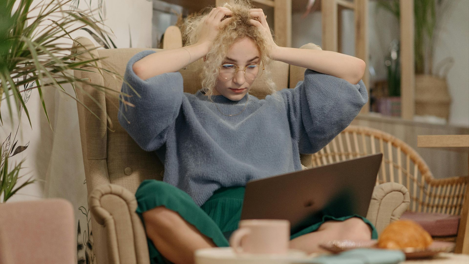 A young woman looking stressed while using her laptop indoors, sitting in an armchair with hands on her head.