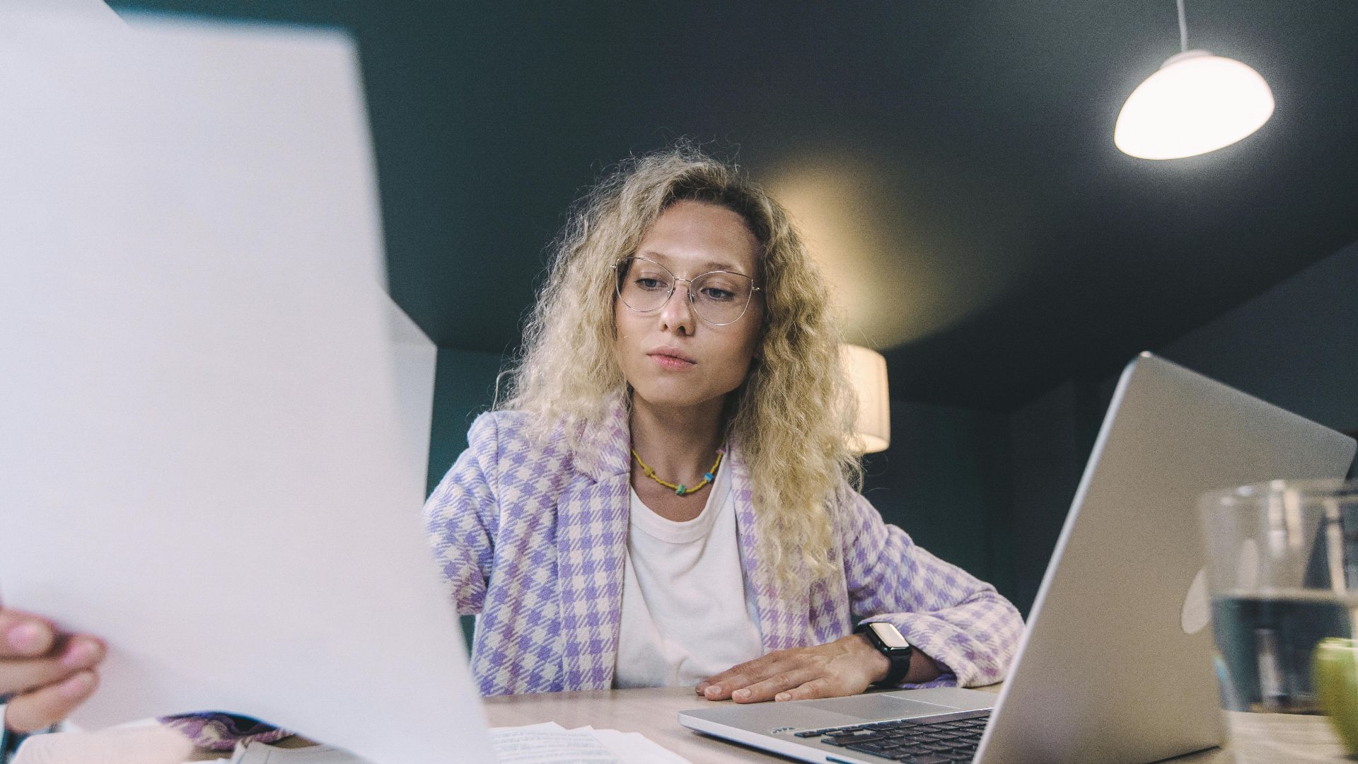 Woman in an office environment reviewing documents with focus, surrounded by technology.