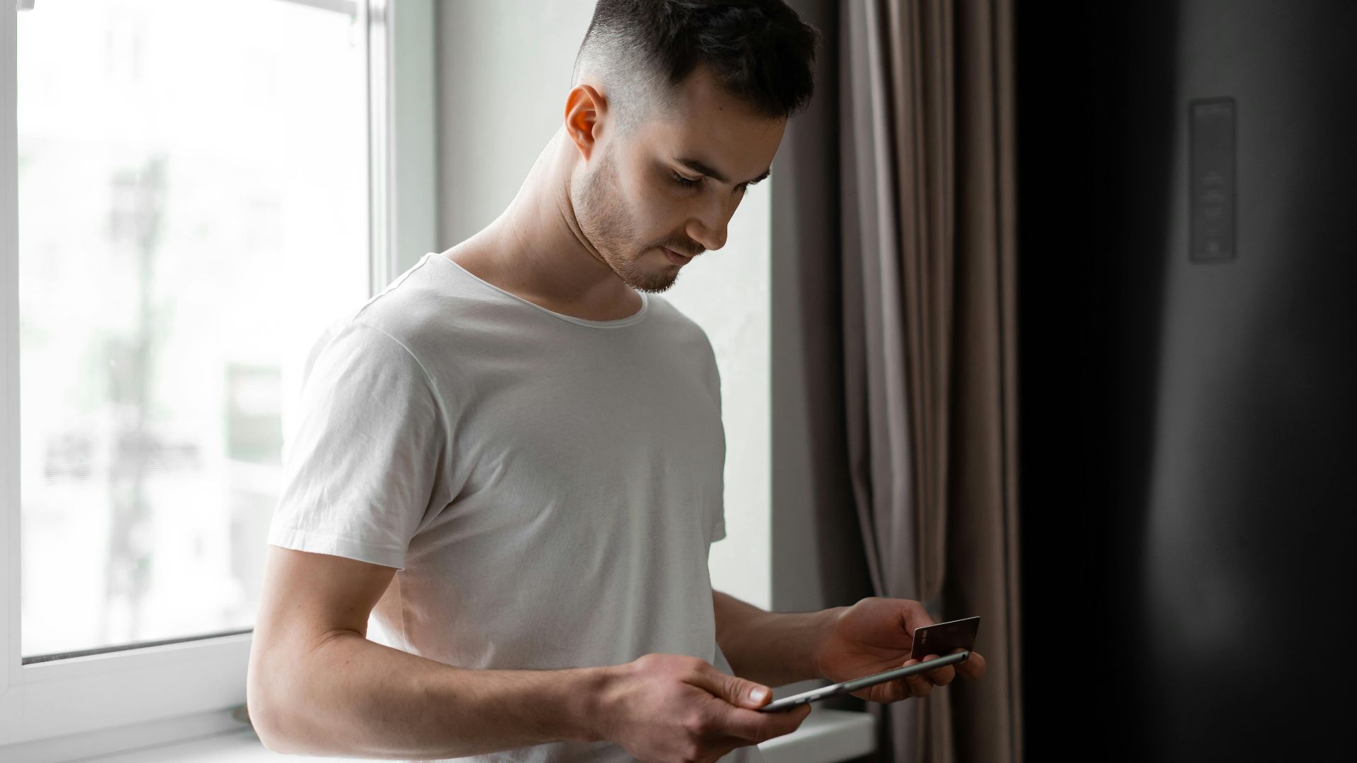 Young man standing by a window, using tablet and credit card for online shopping indoors.