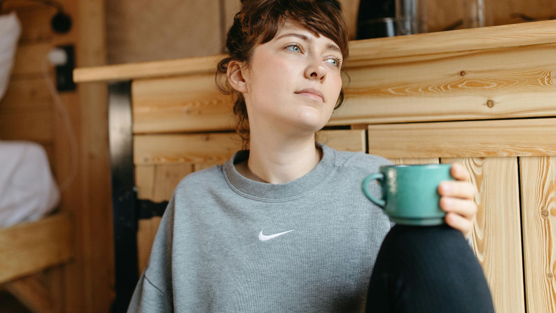 Woman in gray sweater holding a green mug, relaxing in a wooden bedroom setting.