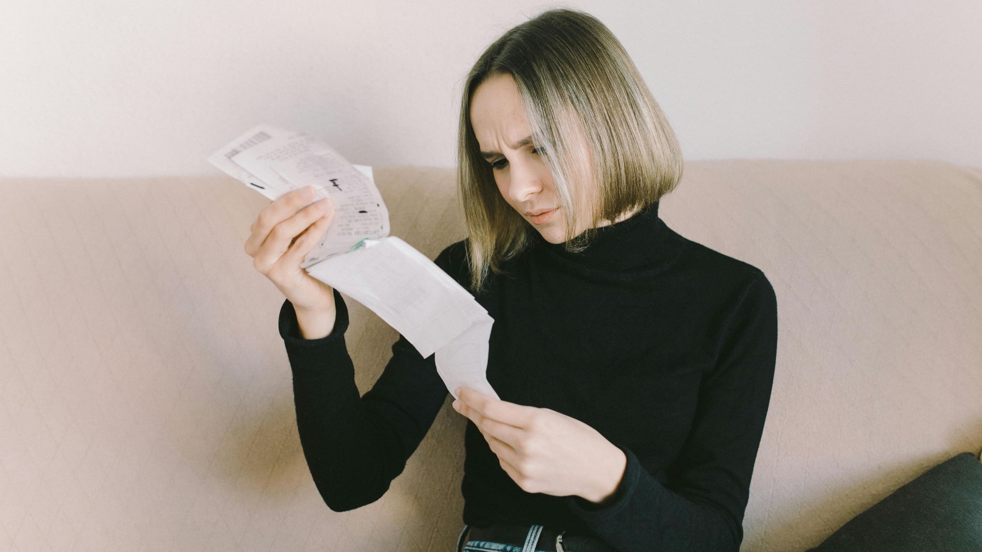 Concerned woman in black sweater examining bills on beige sofa indoors.