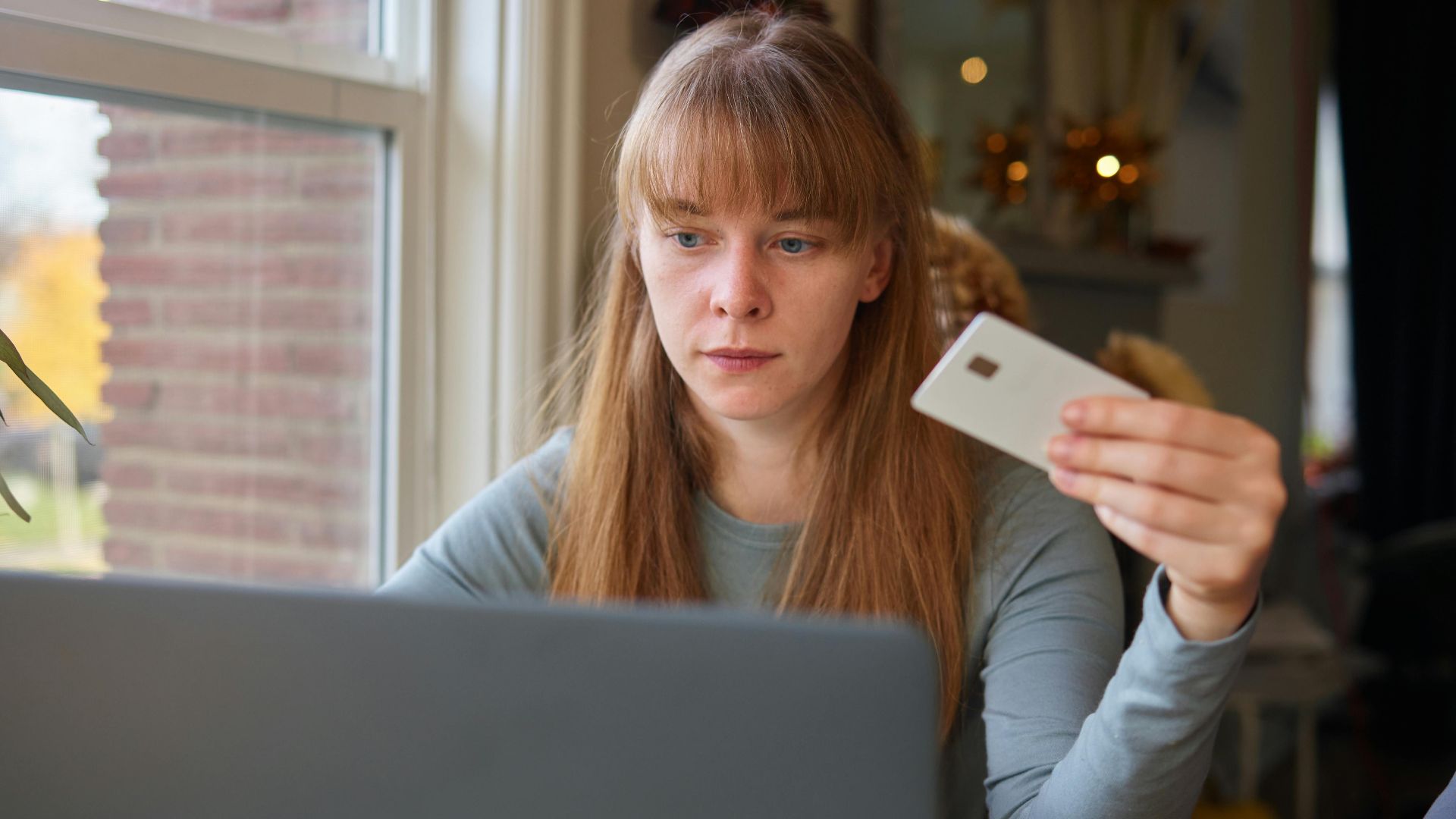 A young woman holds a credit card while using her laptop indoors.