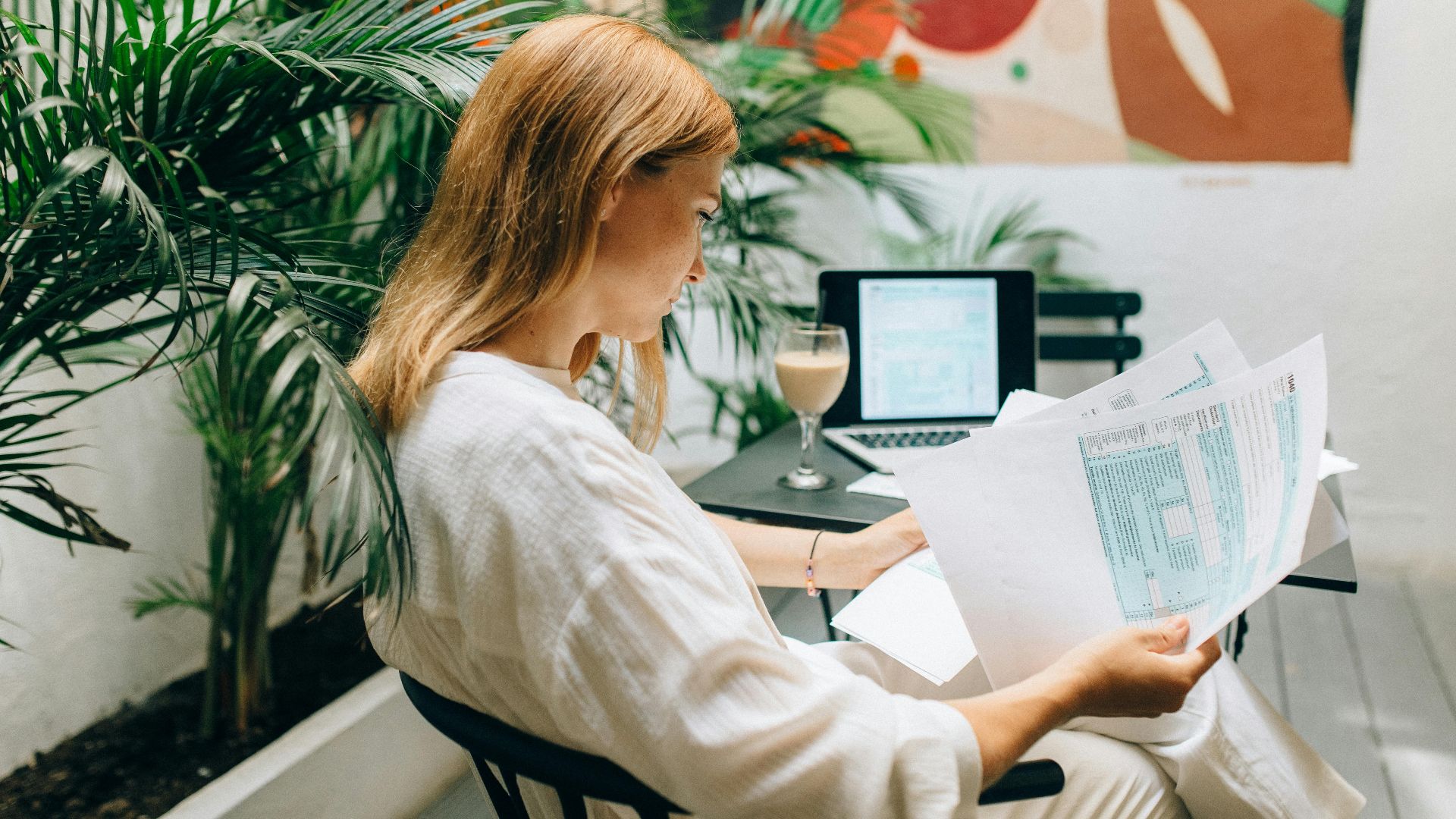 A woman reviews financial documents in an indoor garden space, with a laptop open.