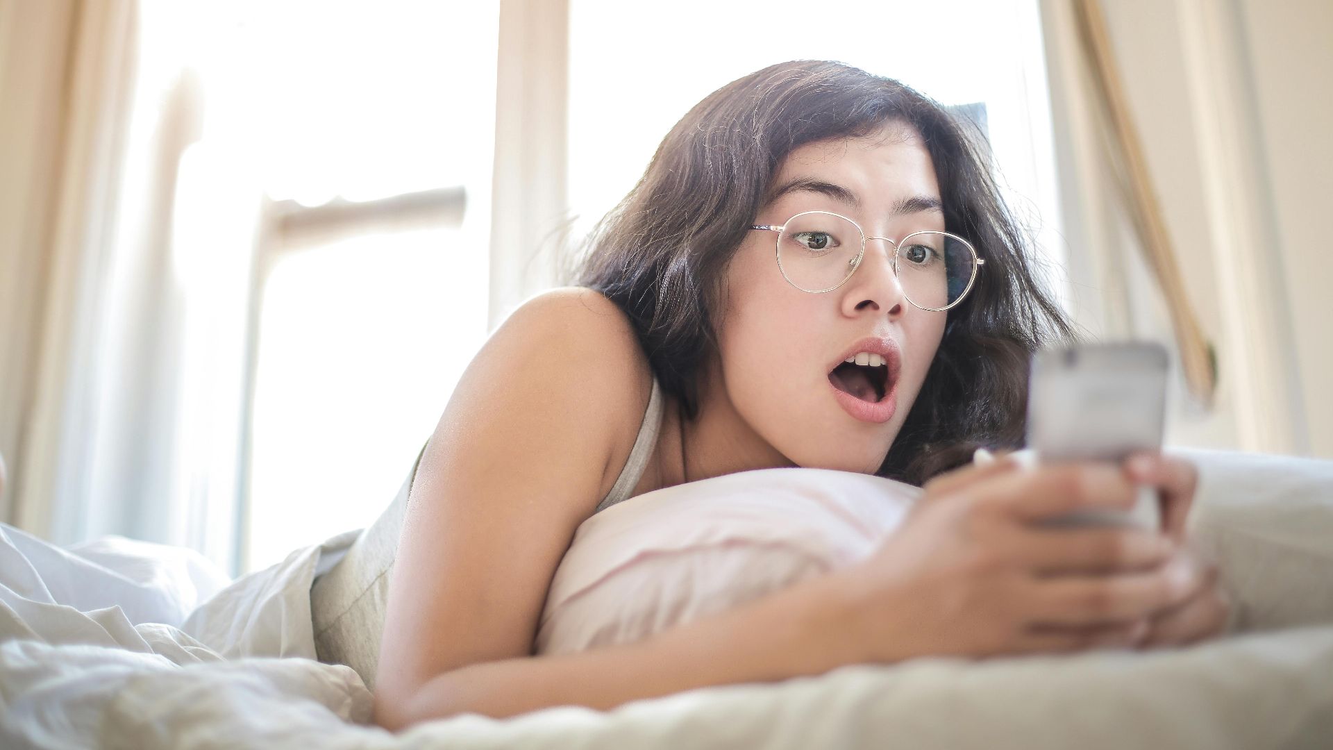 Young woman lying on bed, surprised by something on her smartphone, in a bright bedroom setting.