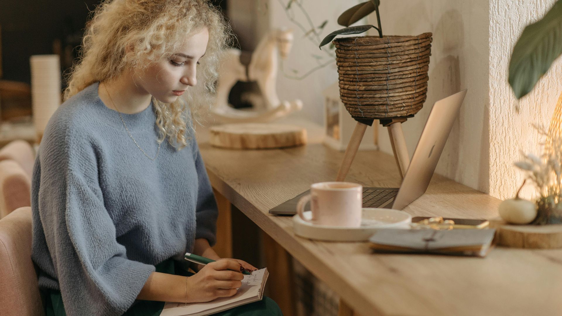 A young woman with curly hair writes in a notebook at a wooden table in a cozy indoor setting.