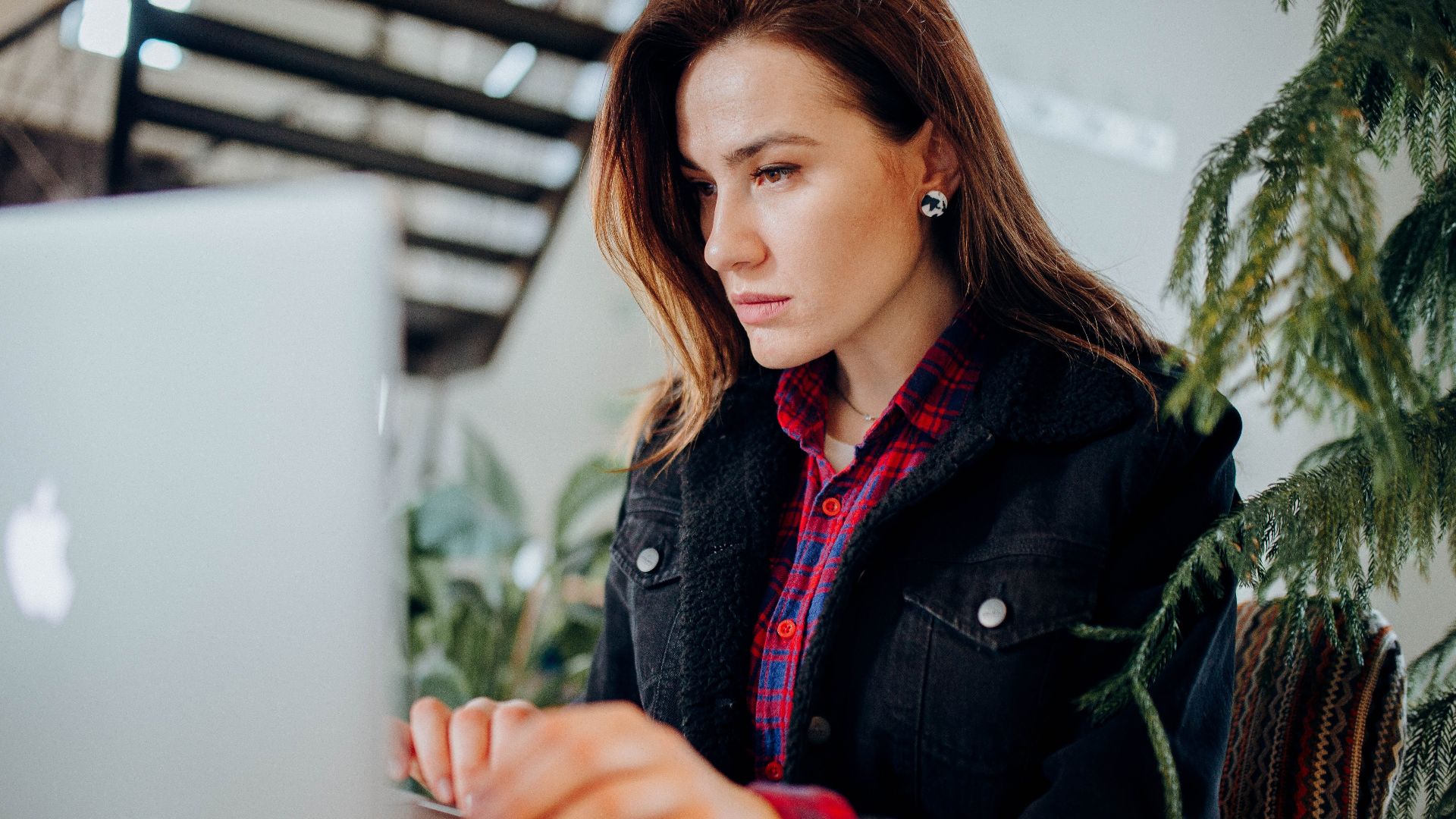 Young woman in plaid shirt intensely working on laptop in a modern environment with greenery.
