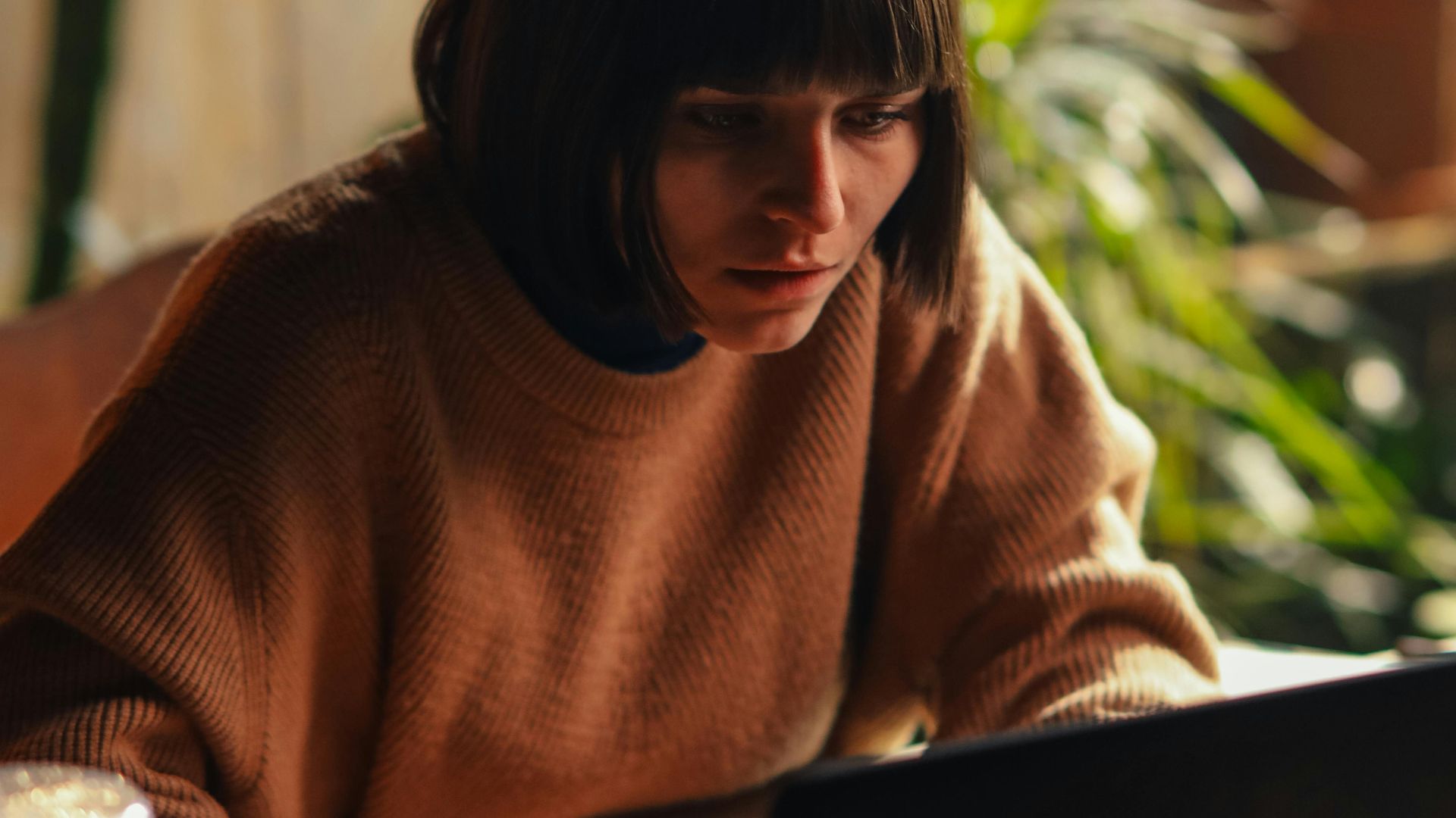 A woman in a cozy sweater concentrates on her laptop in a sunlit room, surrounded by plants.