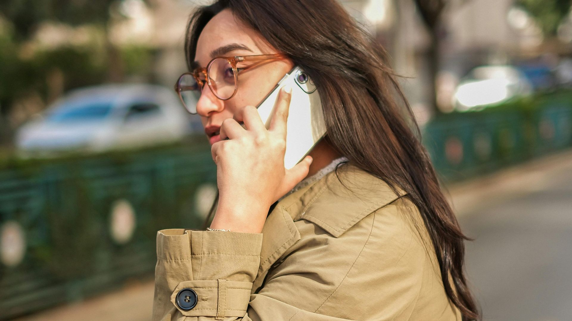 Brunette woman with eyeglasses talking on the phone outdoors in a trench coat.