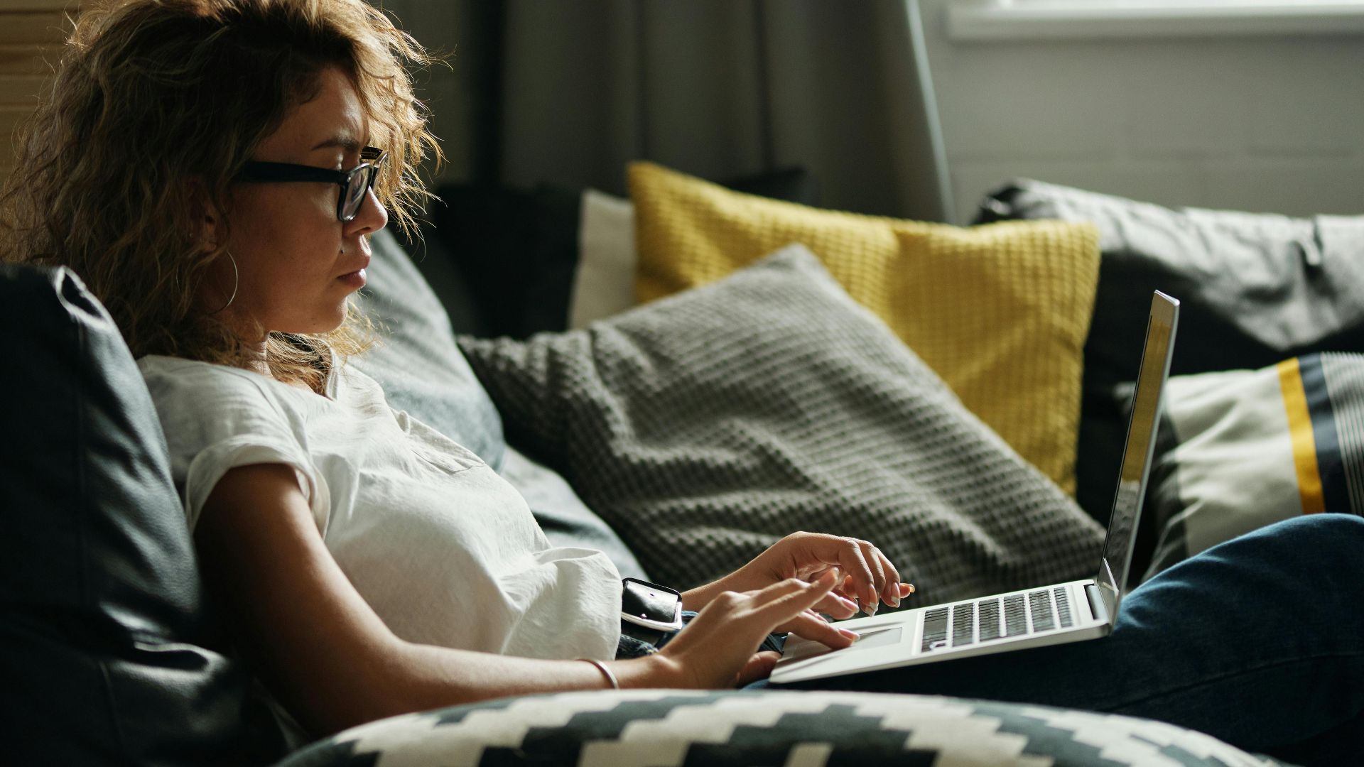 Young woman working on laptop while relaxing on a sofa with pillows, indoors.