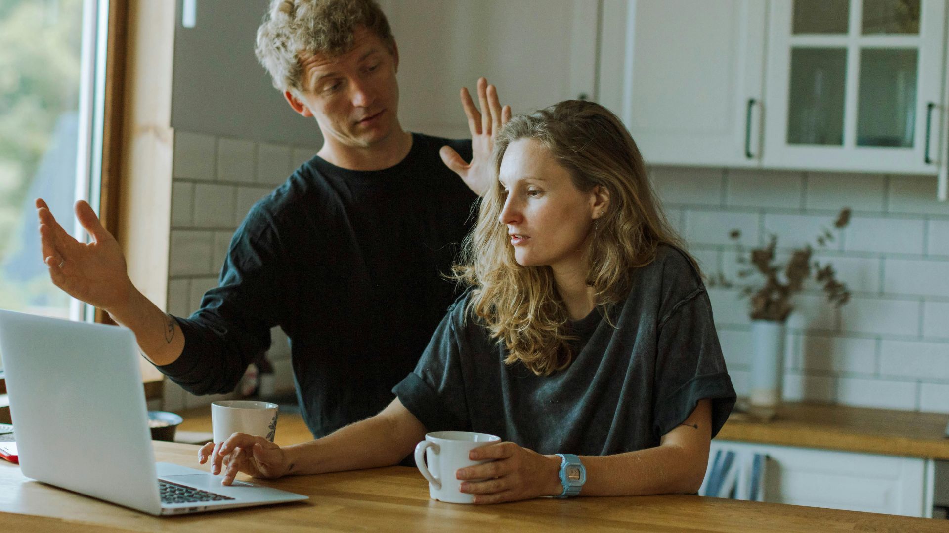 A couple interacting in a bright kitchen using a laptop at a wooden counter.