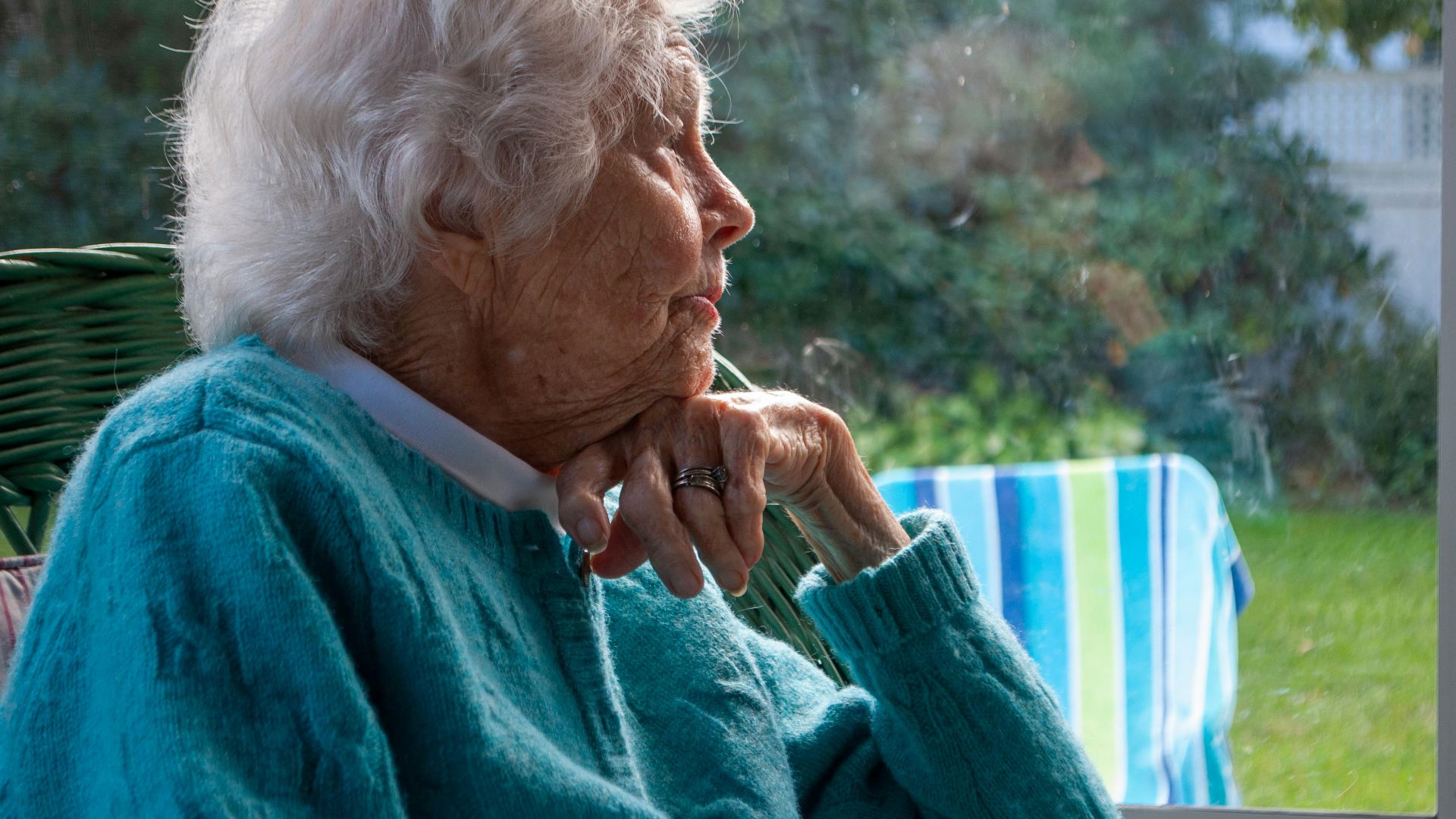 Side view of pensive senior female with white hair in warm sweater leaning on hand and looking away while resting on chair near window at home