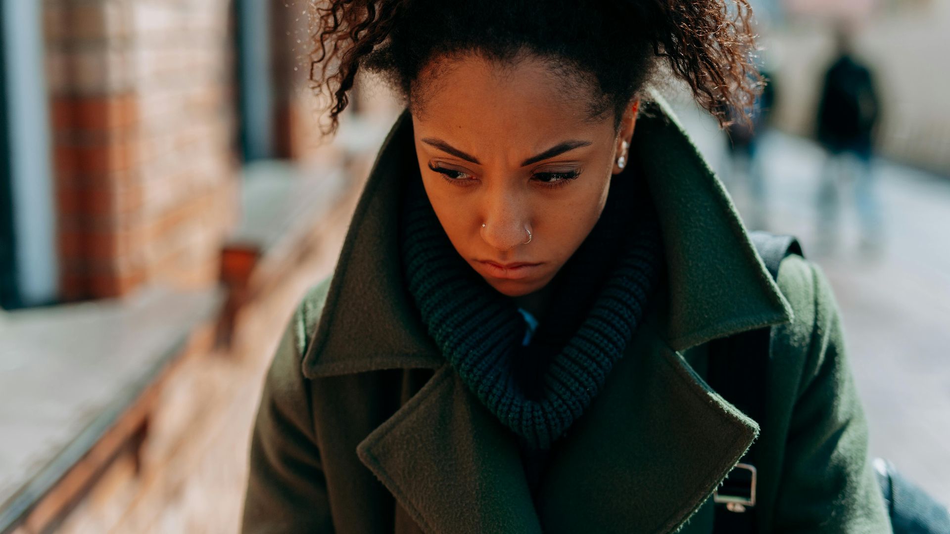 A young woman in a trench coat appears thoughtful and pensive walking down a city street.