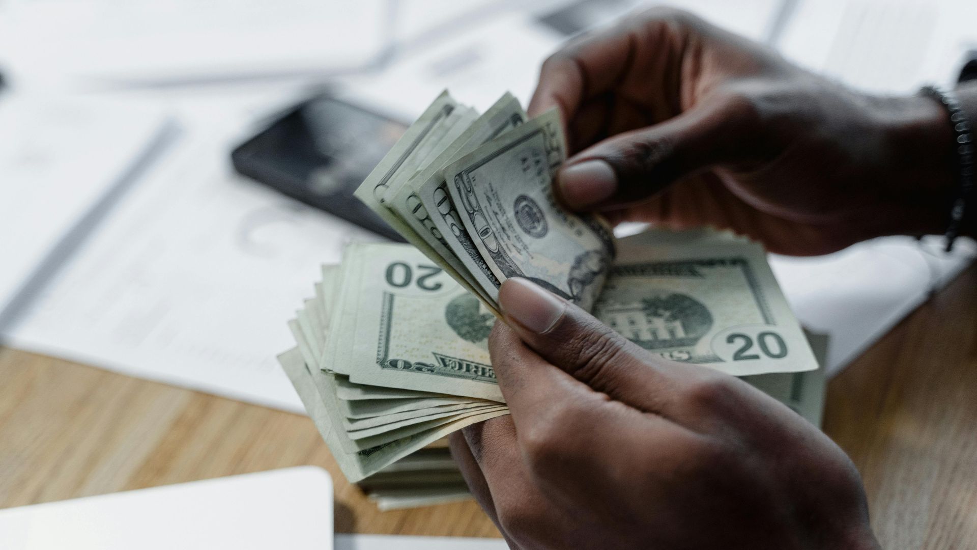 A close-up of hands counting US dollar bills on a wooden table with documents and a smartphone nearby.