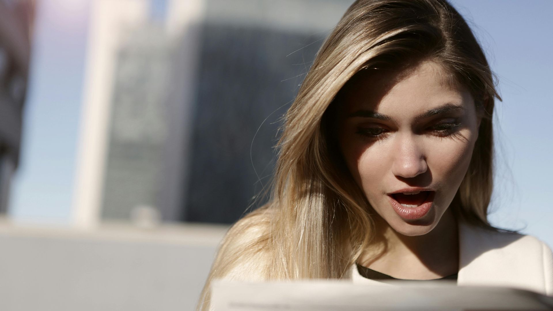 A young woman with blonde hair expresses surprise while reading a newspaper outdoors in daylight.