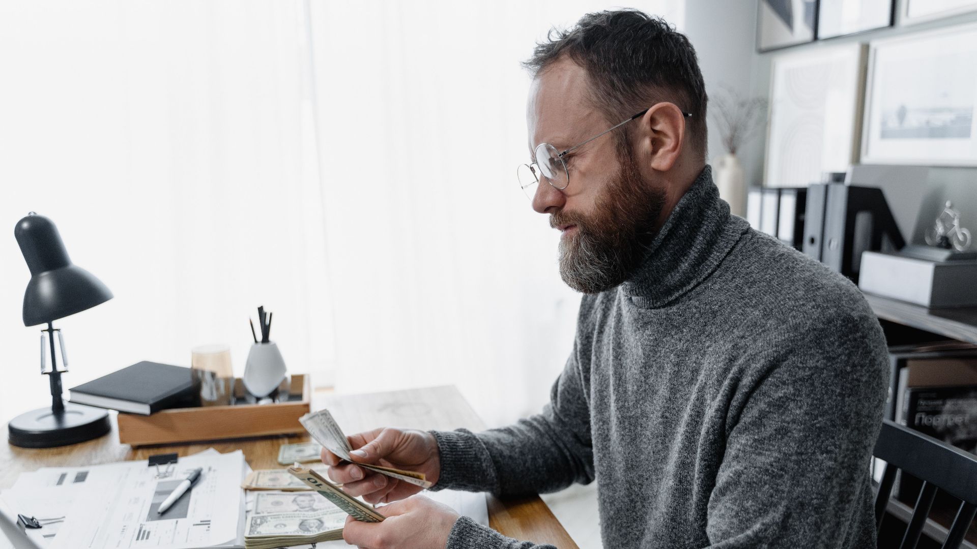 Businessman wearing glasses counting cash at office desk with documents and smartphone.