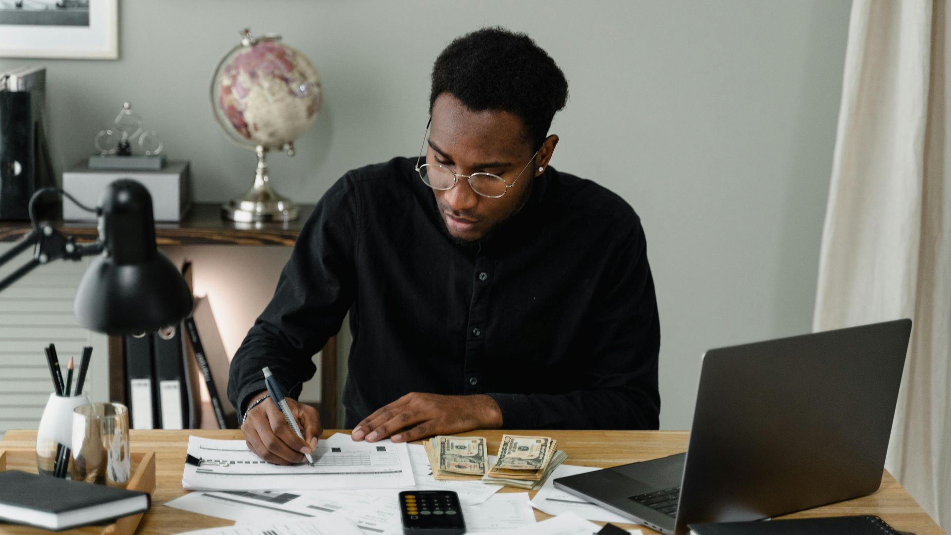 An adult man calculates expenses, using a laptop and documents at a desk in a home office setting.