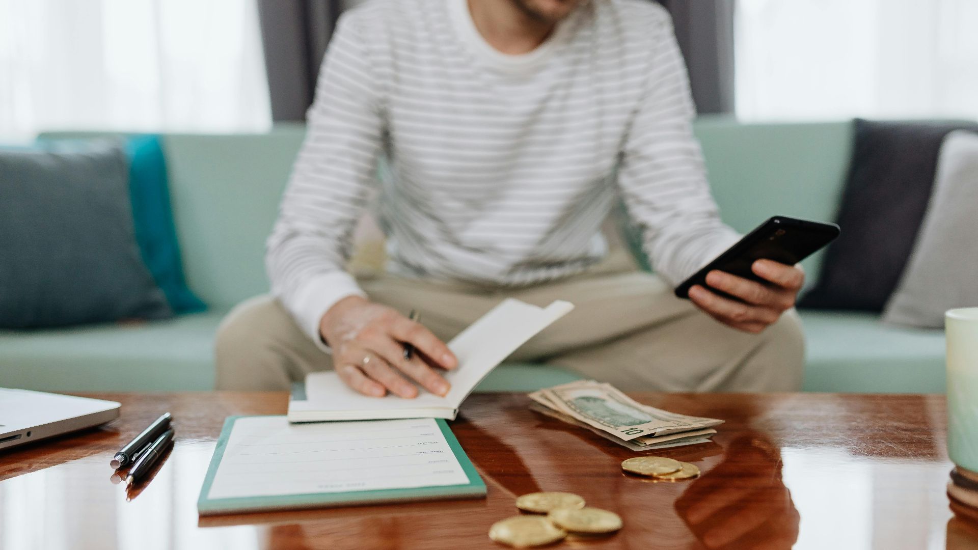 Adult man sits on sofa managing finances with cash, smartphone, and notebook.