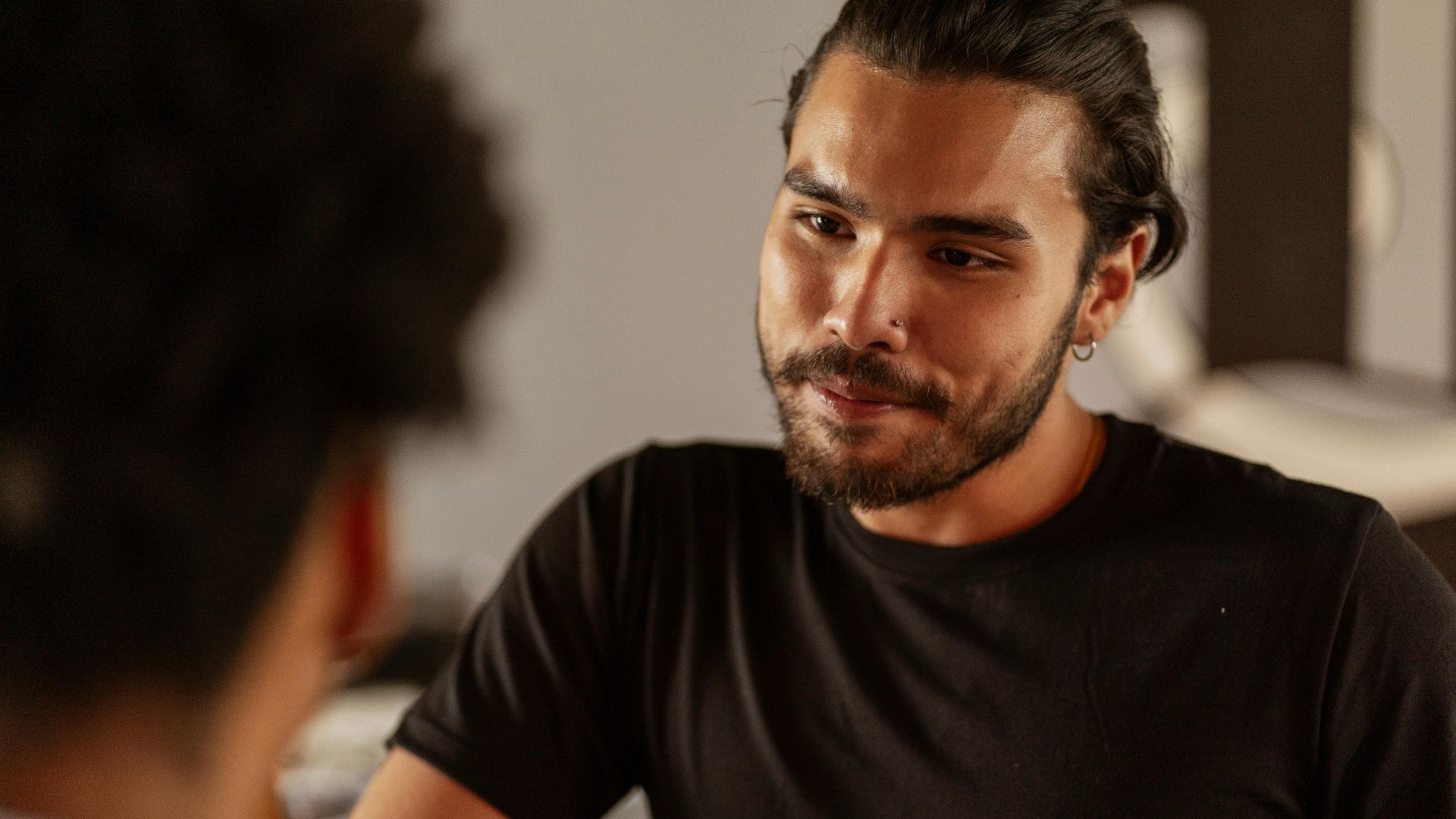A bearded man attentively engaged in a conversation indoors, wearing a black shirt.