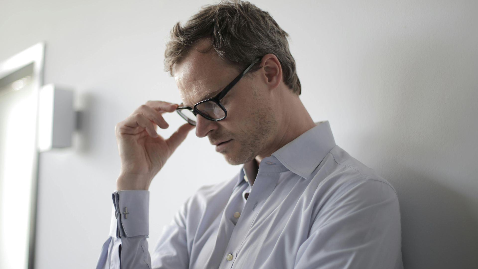 An adult man in a shirt and glasses appears stressed while leaning against a wall indoors.