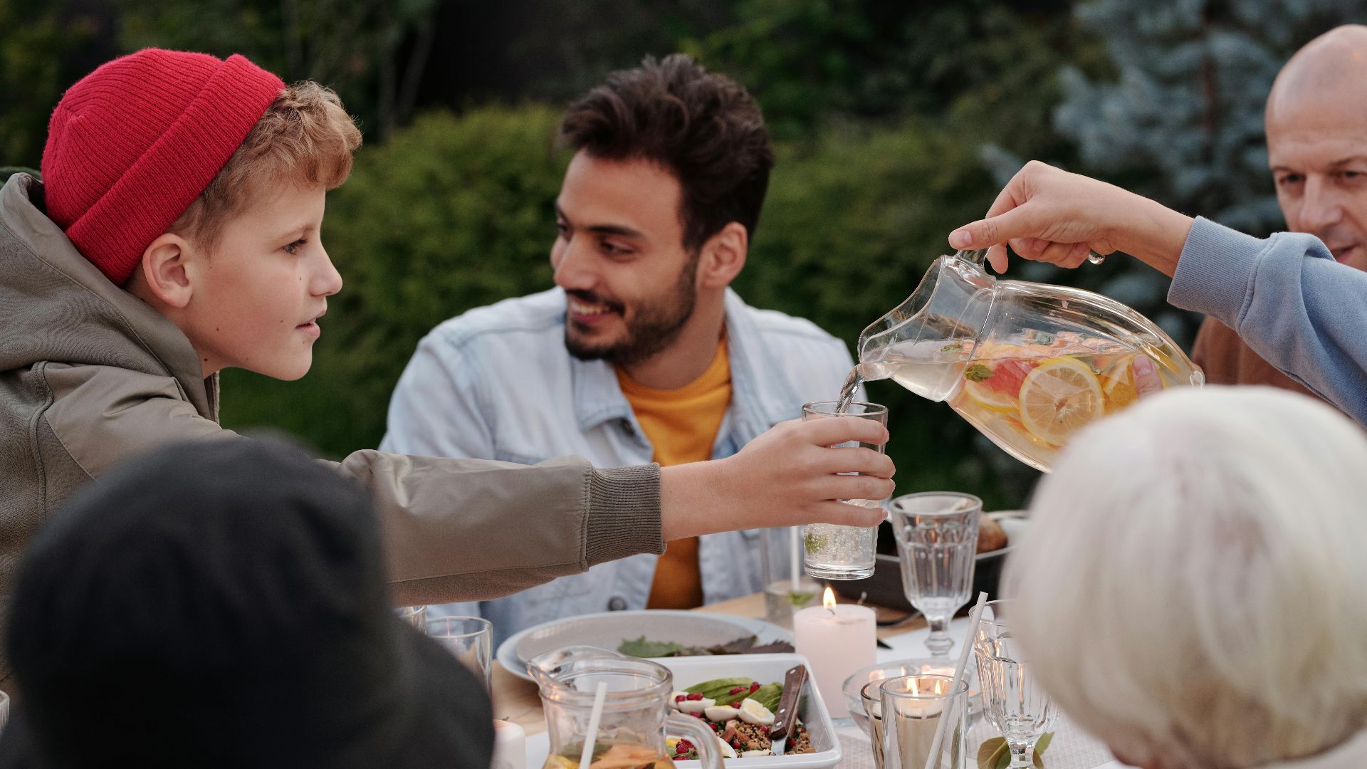 Faceless female pouring fruit beverage into glass of boy in warm cloth while cheerful multiethnic family having dinner outdoors