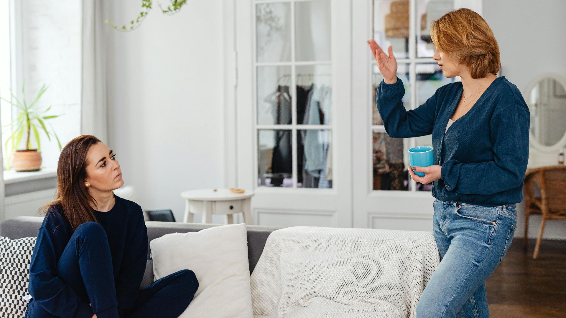Two women engage in a friendly chat in a stylish living room, one sitting and one standing with a mug.