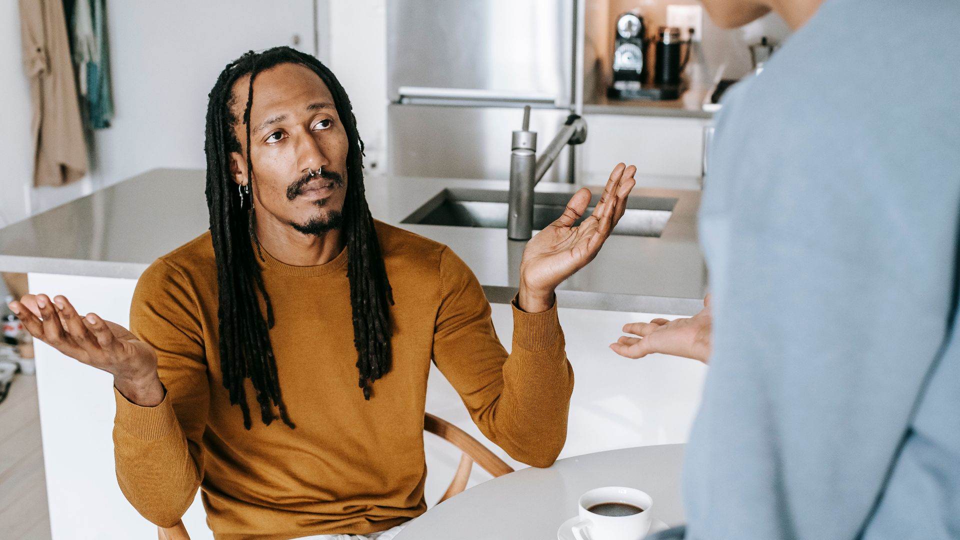 Black male sitting at table and arguing with standing near table female in casual outfit in light kitchen
