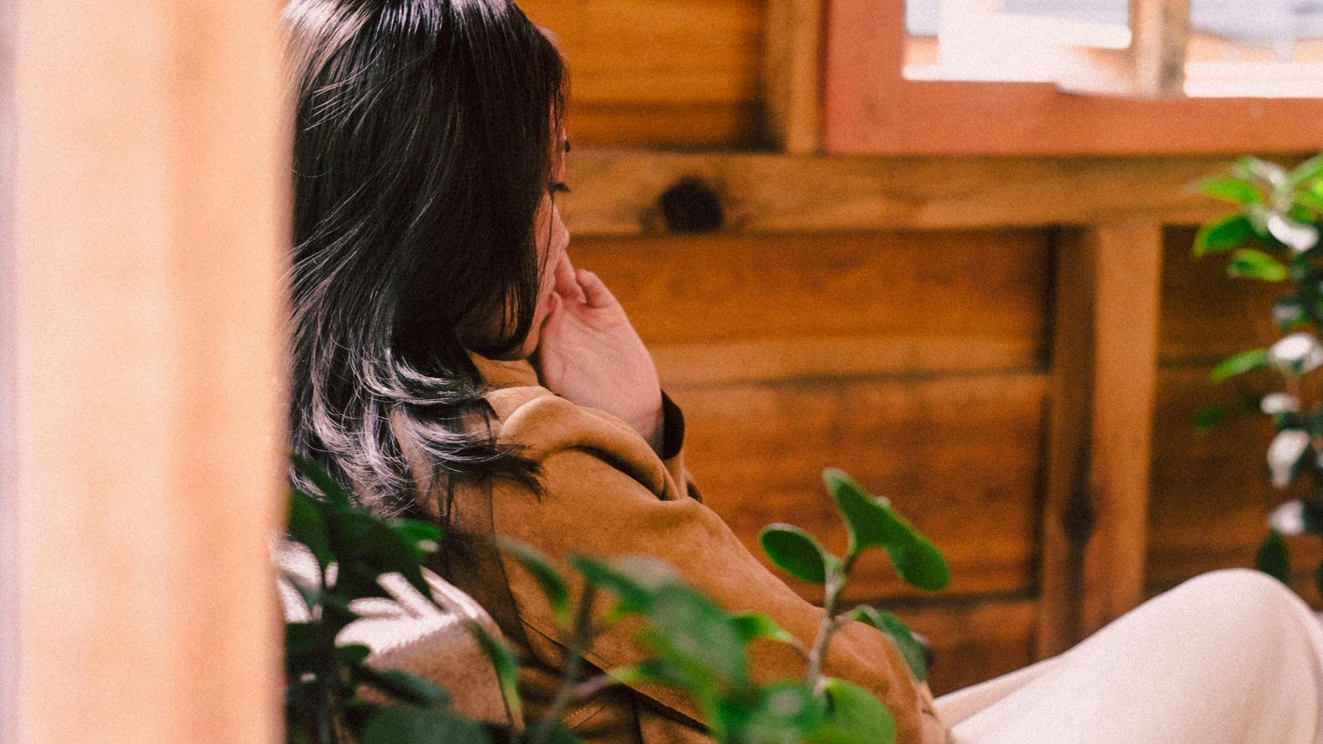 Asian woman sitting indoors in a wooden house, surrounded by greenery, reflecting tranquility.