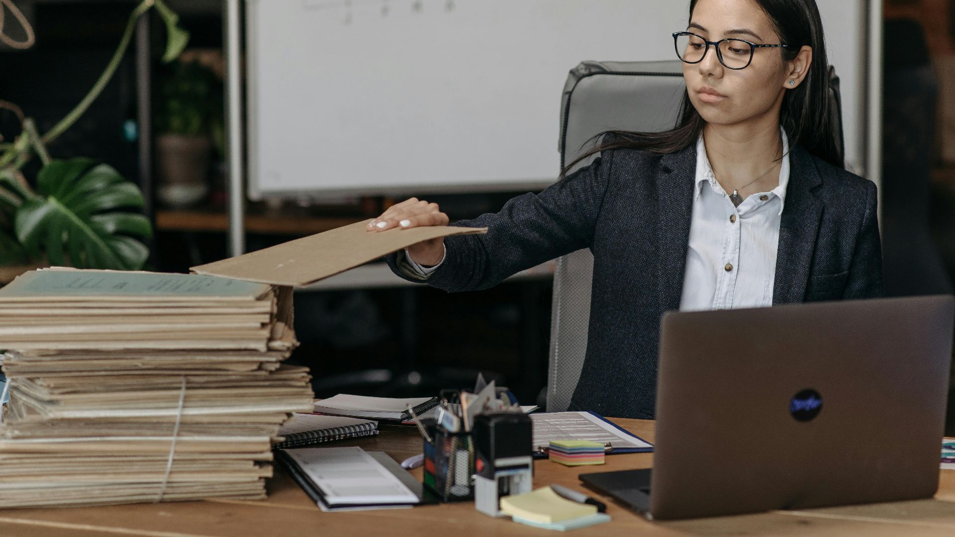 A businesswoman in a modern office organizing a stack of files beside a laptop.