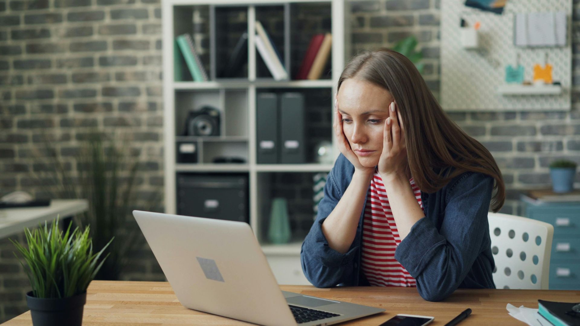 A woman appears stressed while working on a laptop in her modern office.