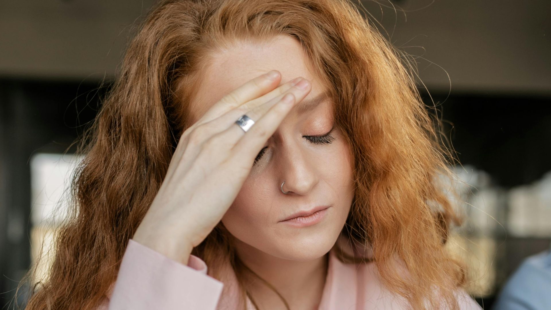 Stressed woman with red hair in pink shirt facing work pressure. Indoor office scene.