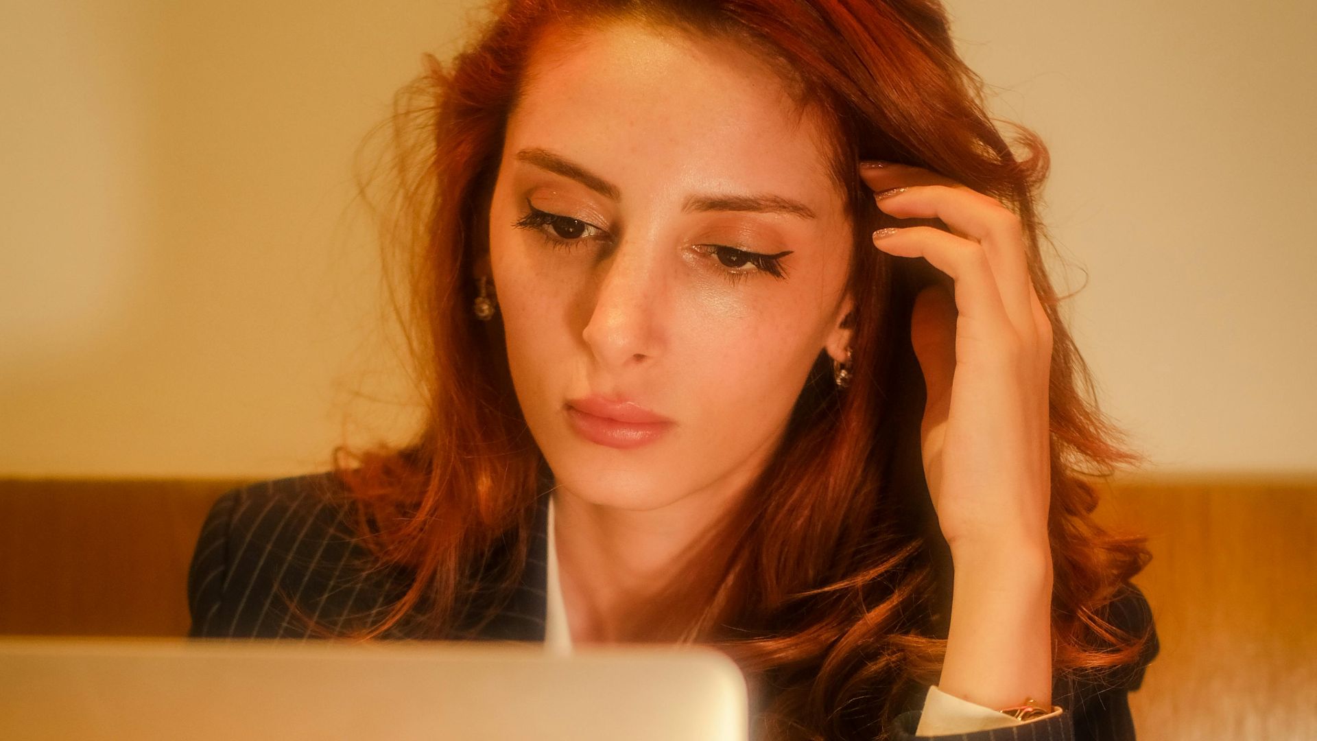 Red-haired woman in a business suit working intently on a laptop in an office setting.