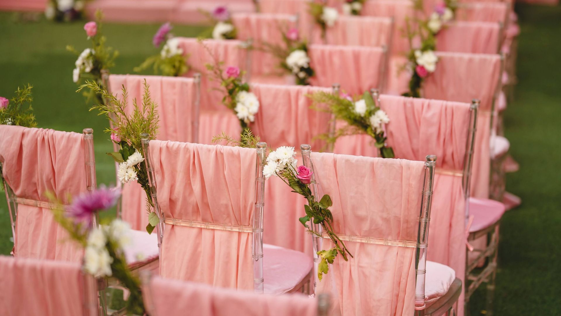 Beautifully arranged wedding ceremony seats with pink covers and floral decor on a green lawn.
