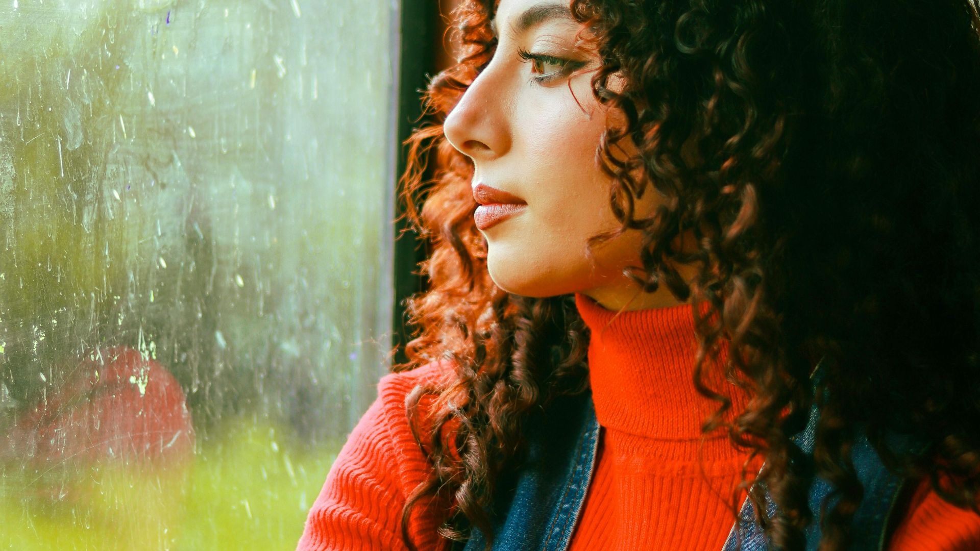 A woman with curly hair in a red sweater sits gazing out a raindrop-covered window.