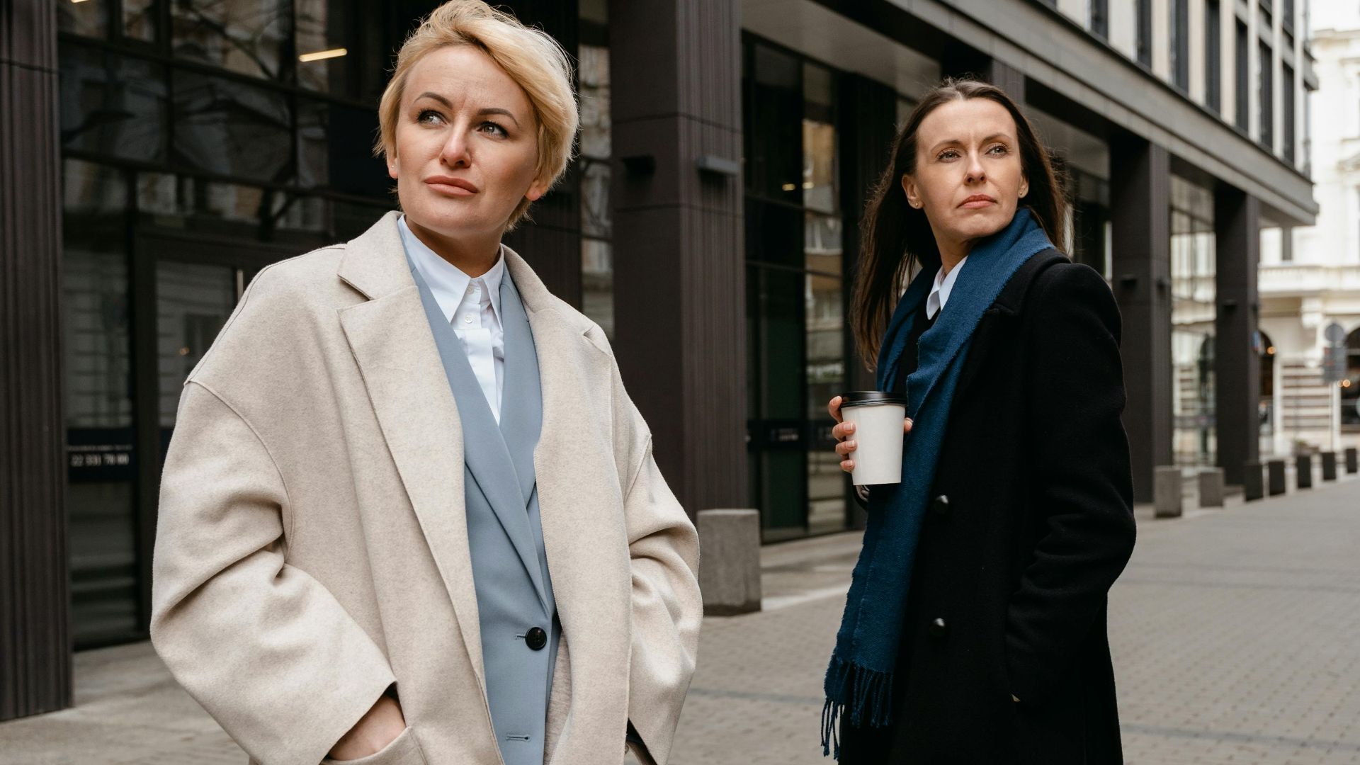 Two professional women in coats standing on a city street, presenting a confident pose.