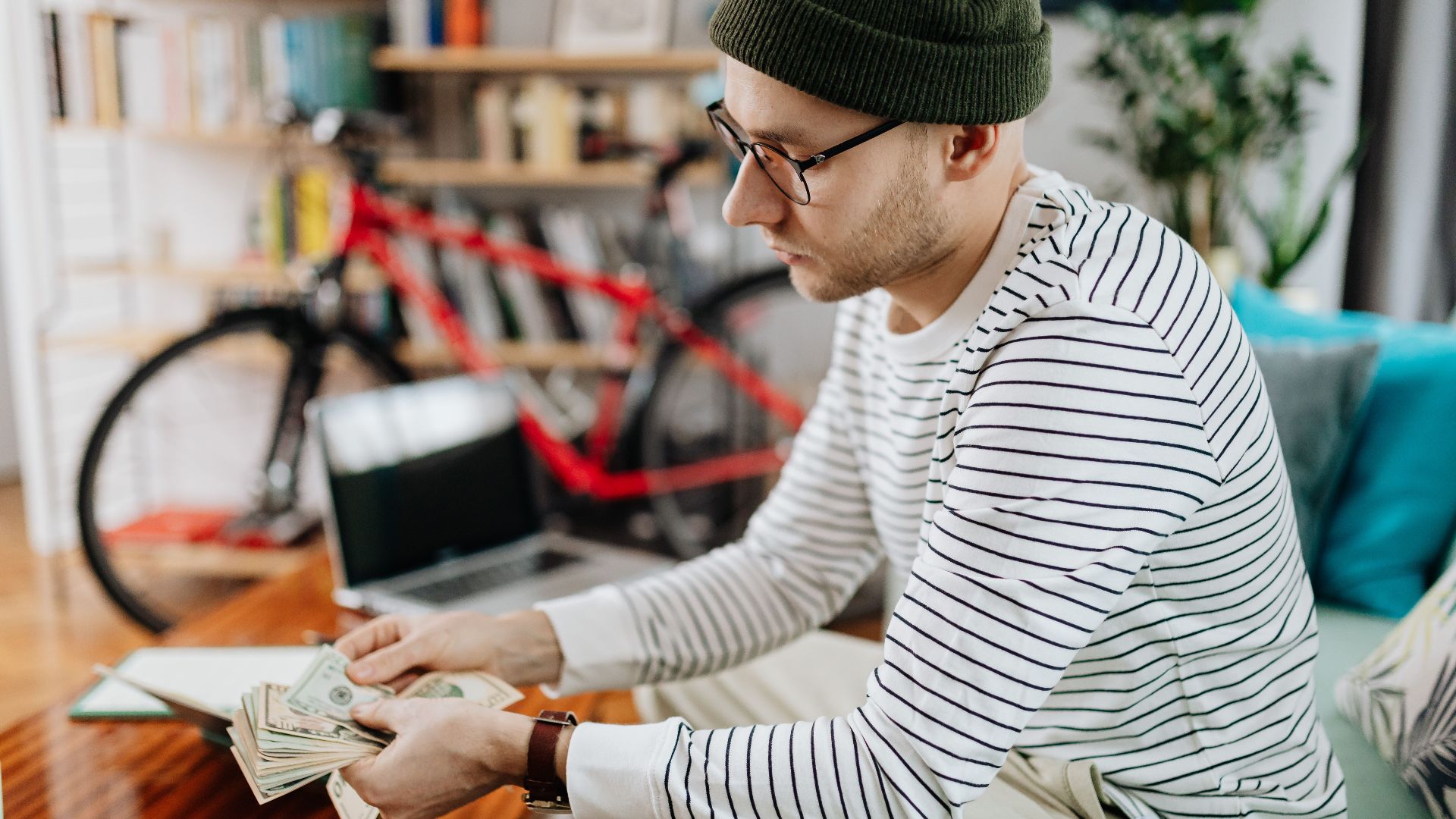 Man in striped shirt counting cash indoors, with a bicycle in the background.
