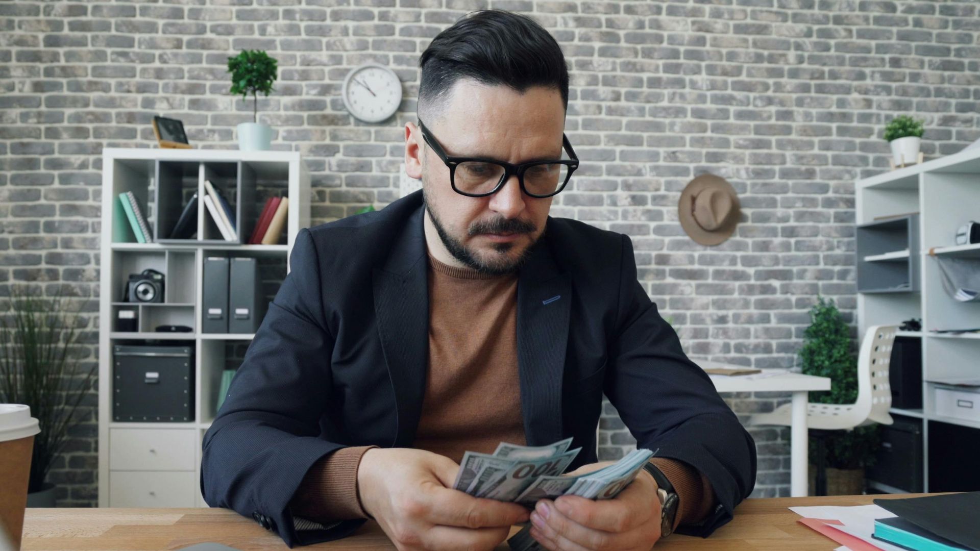 Businessman counting cash at a desk in a modern office setting with a laptop and documents.