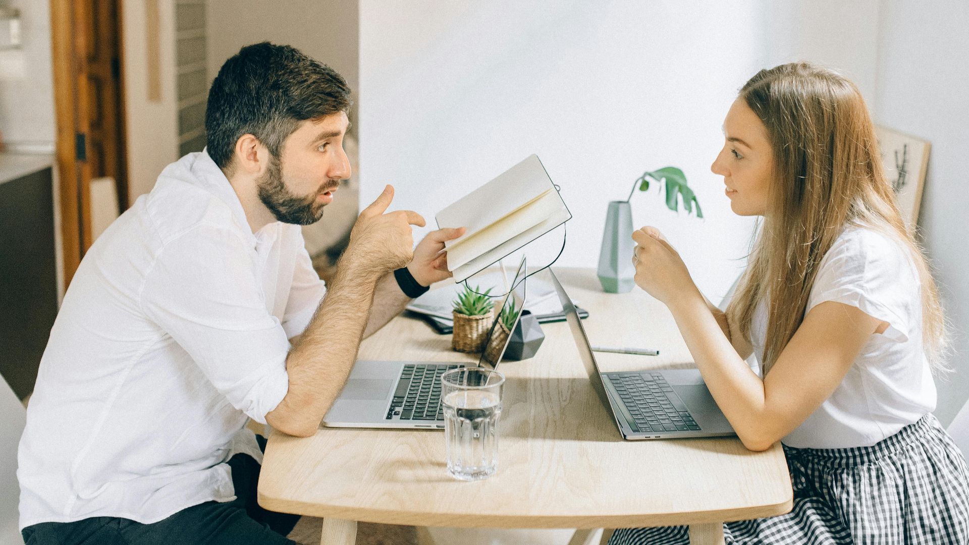 A man and woman working from home on laptops, engaged in discussion at a wooden table.