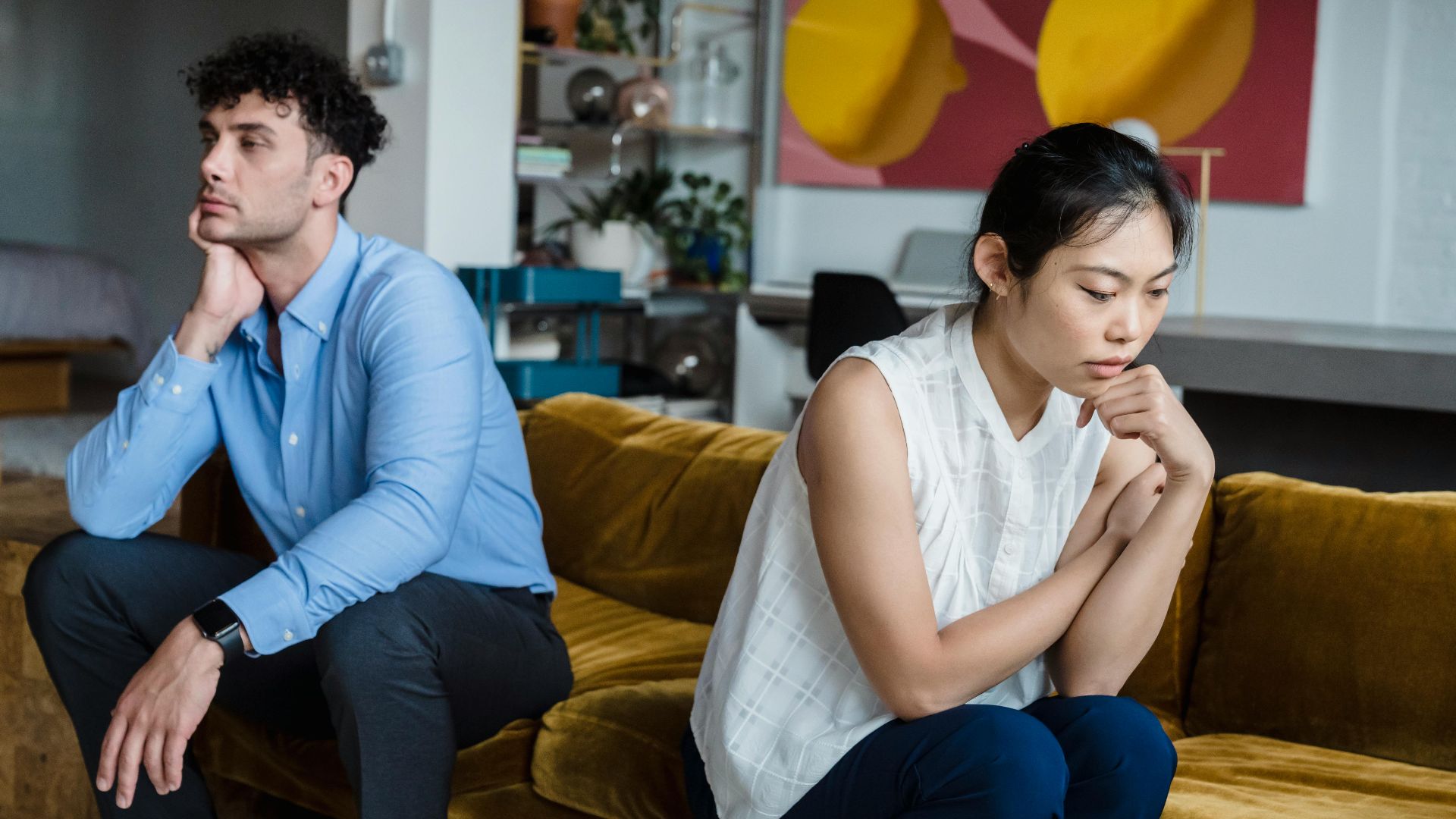 A couple sits indoors on a sofa, both appearing thoughtful and contemplative.