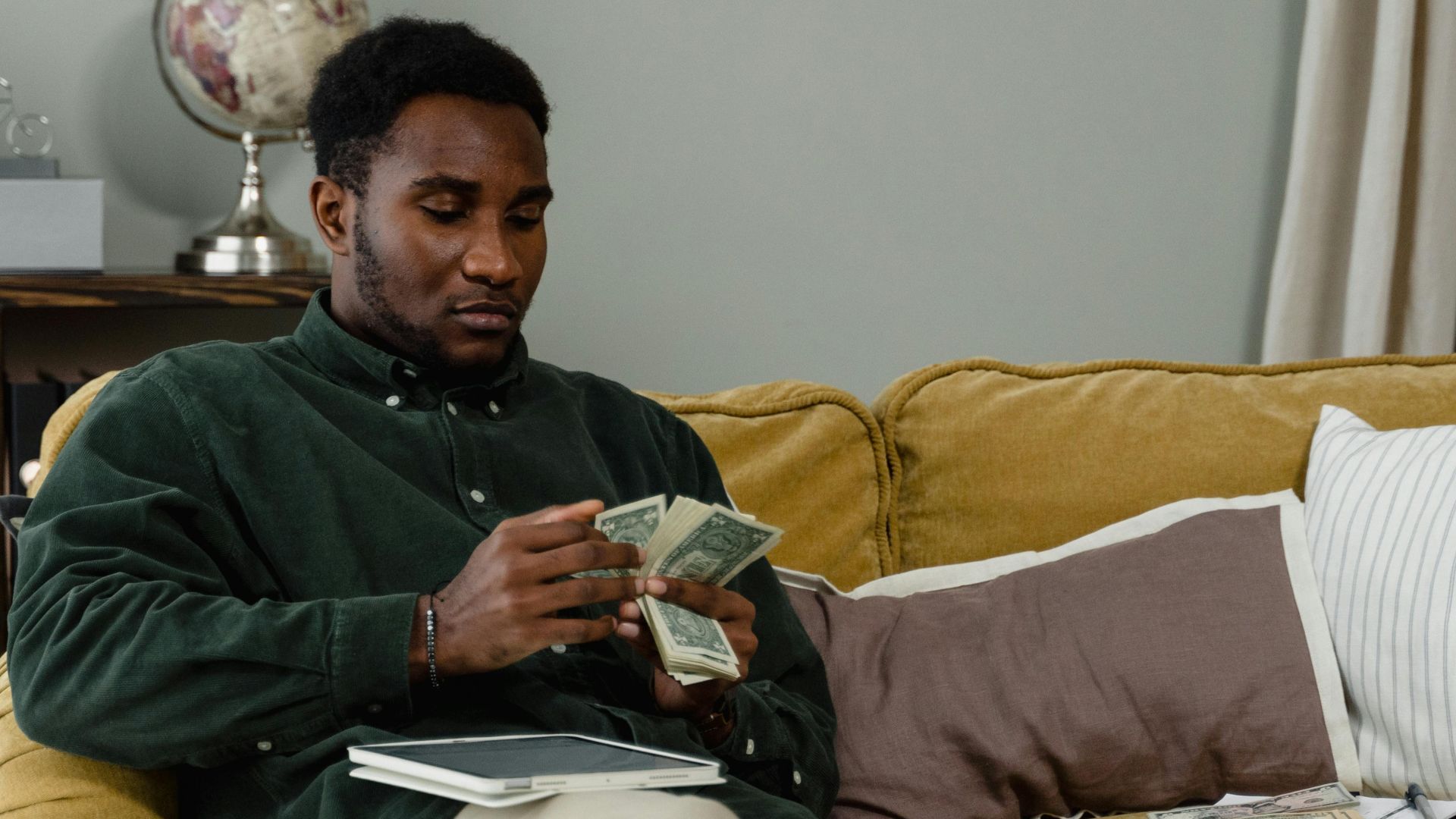 A man sitting on a couch counting cash notes in a cozy indoor setting with a globe in the background.