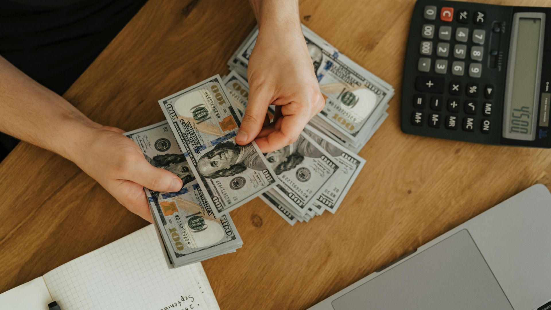 Close-up of hands counting dollar bills with calculator, notebook, and laptop on wooden table.