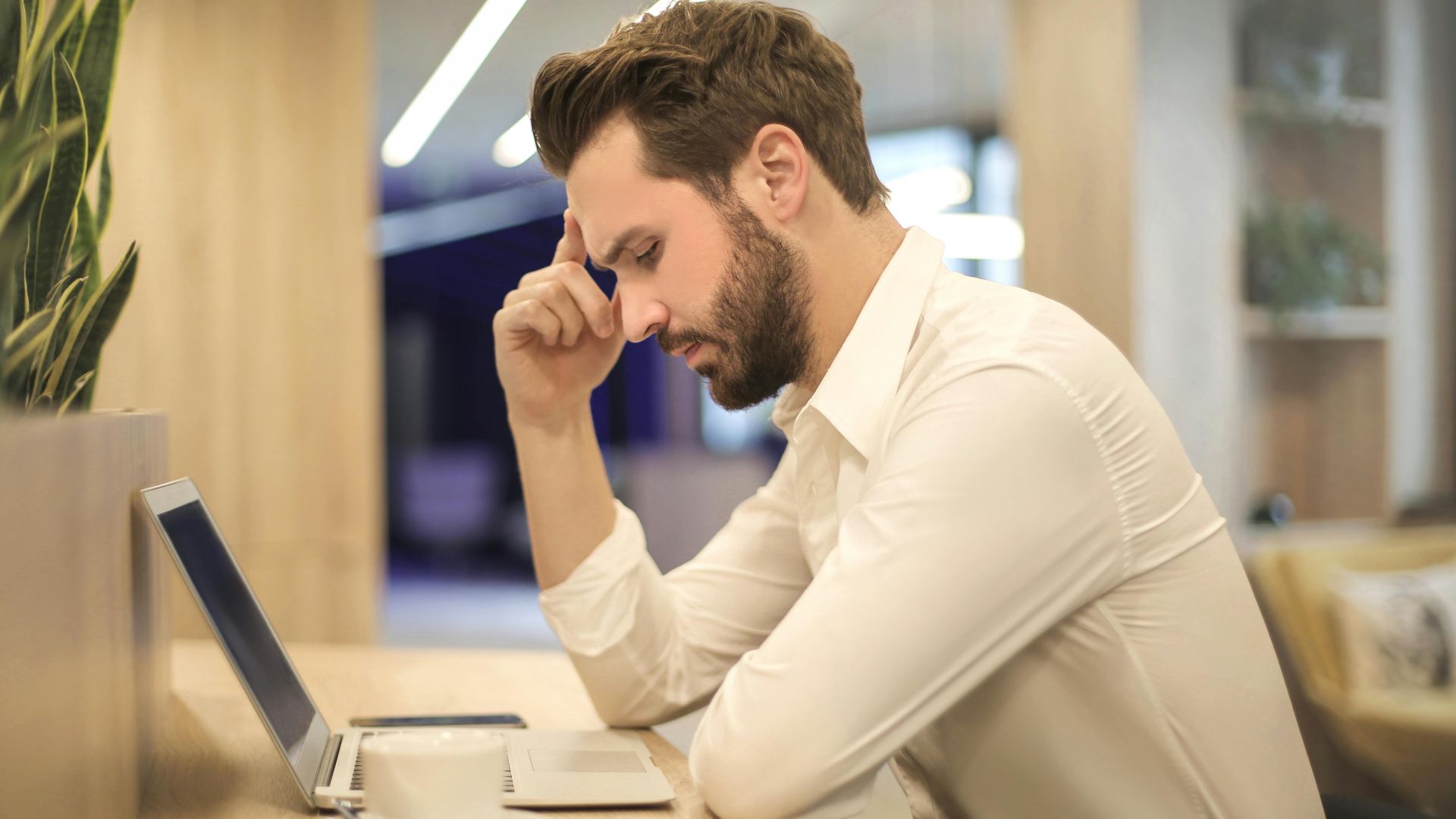 A thoughtful man in a shirt works on his laptop at a modern indoor office space.