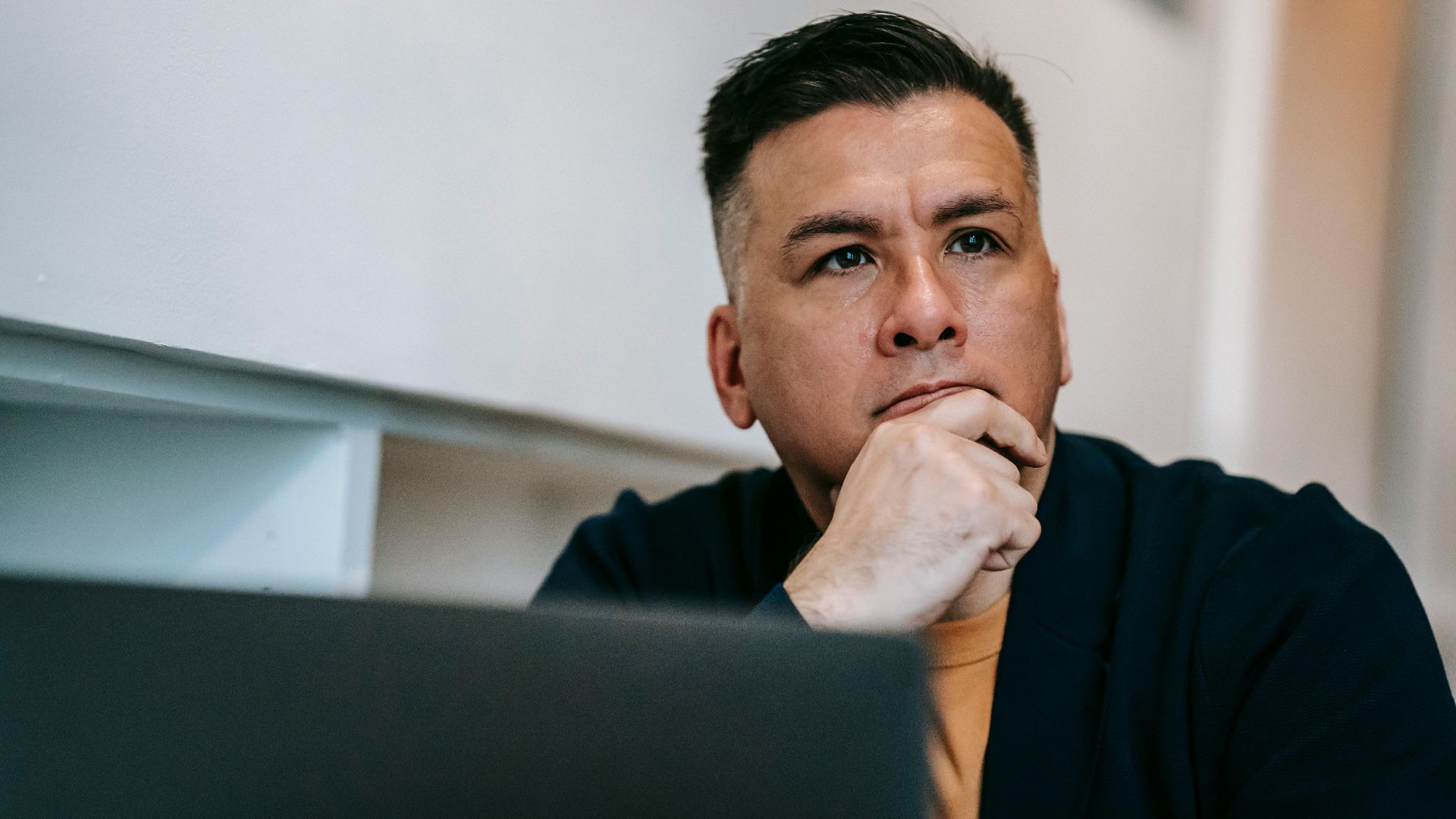 Focused man in home office pondering work on a laptop, expressing deep concentration and thought.