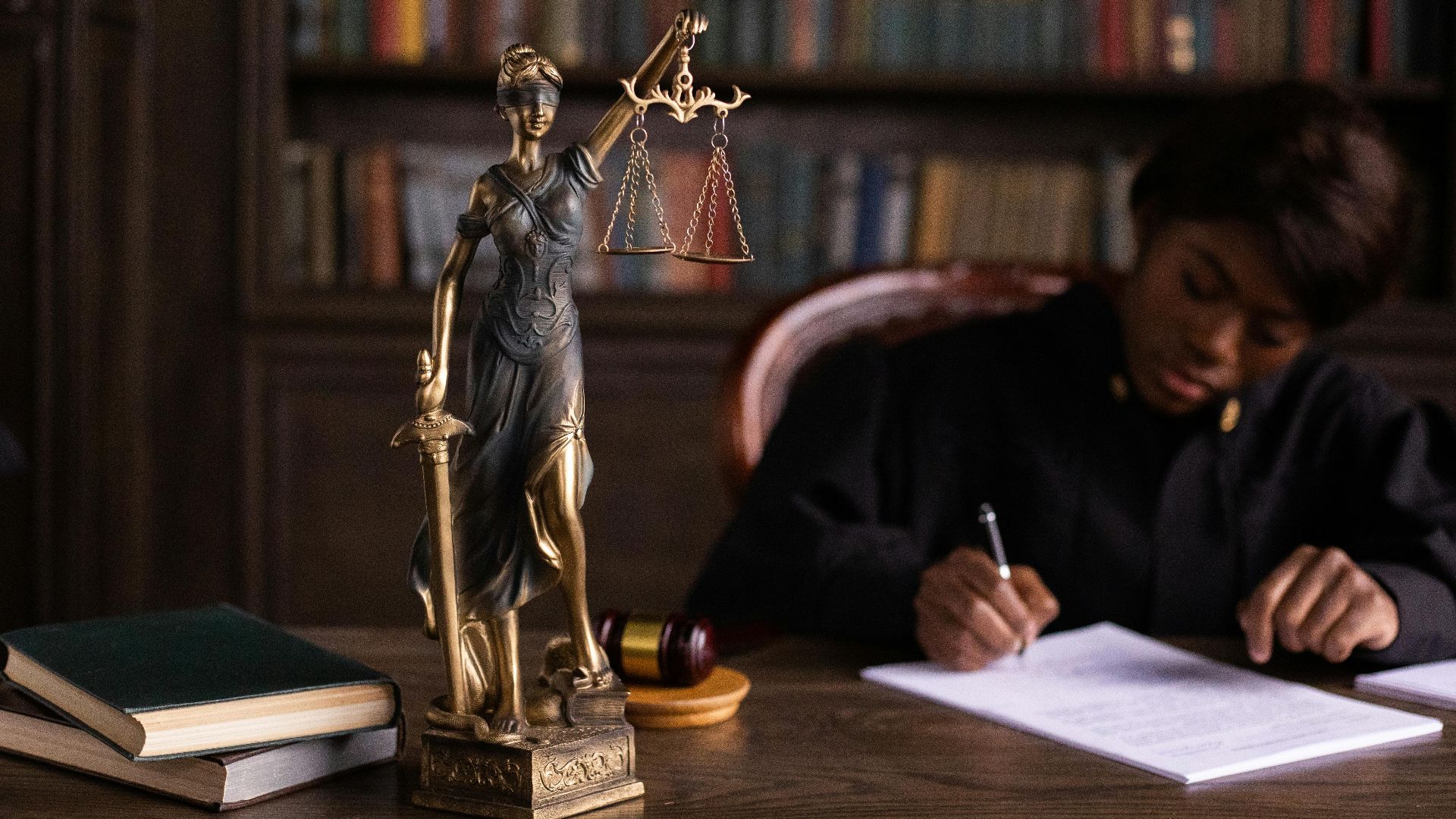 A focused judge writing on documents beside a Lady Justice statue in an office.