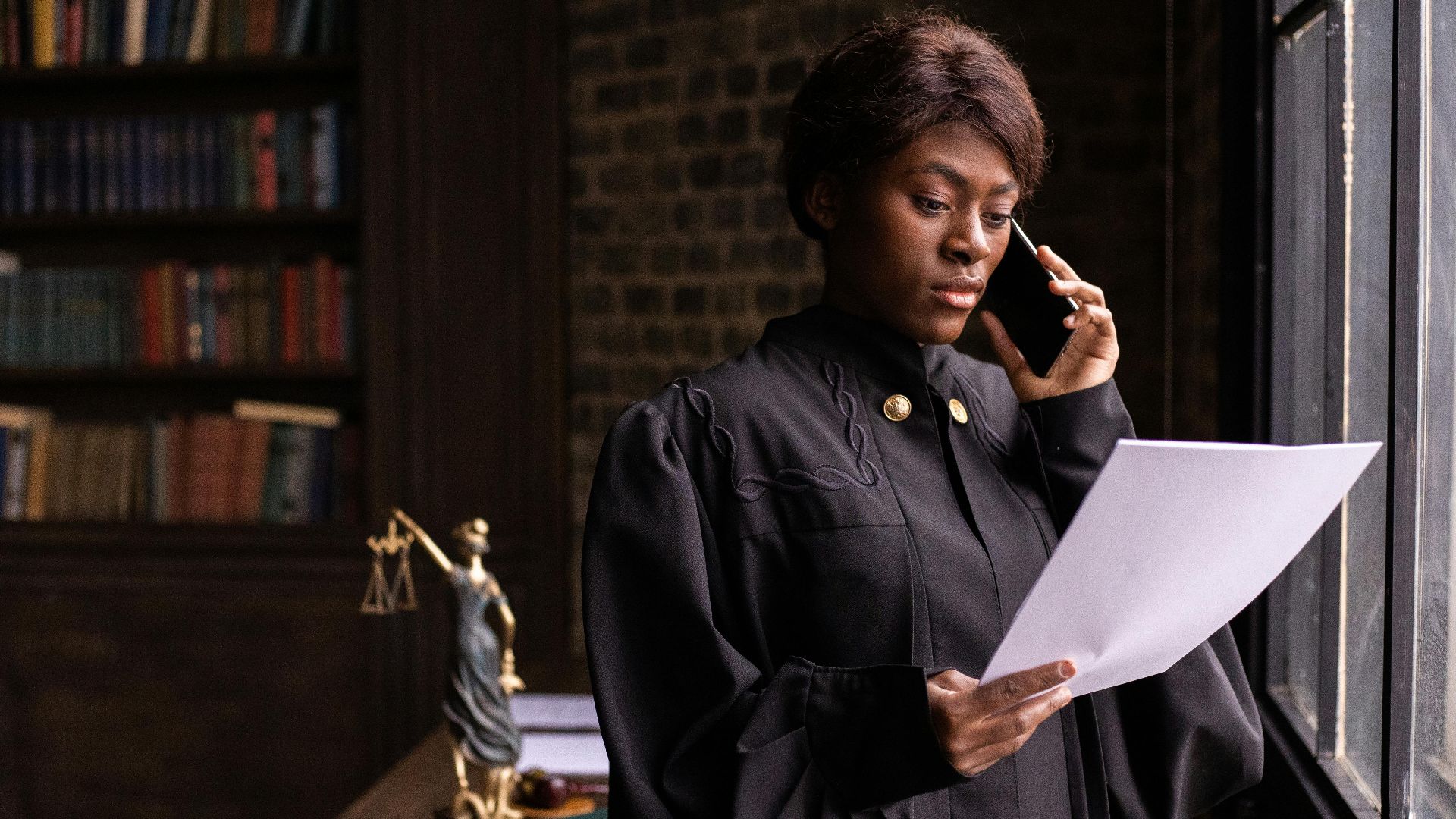 African American judge in robe, reading document and talking on phone by the office window.