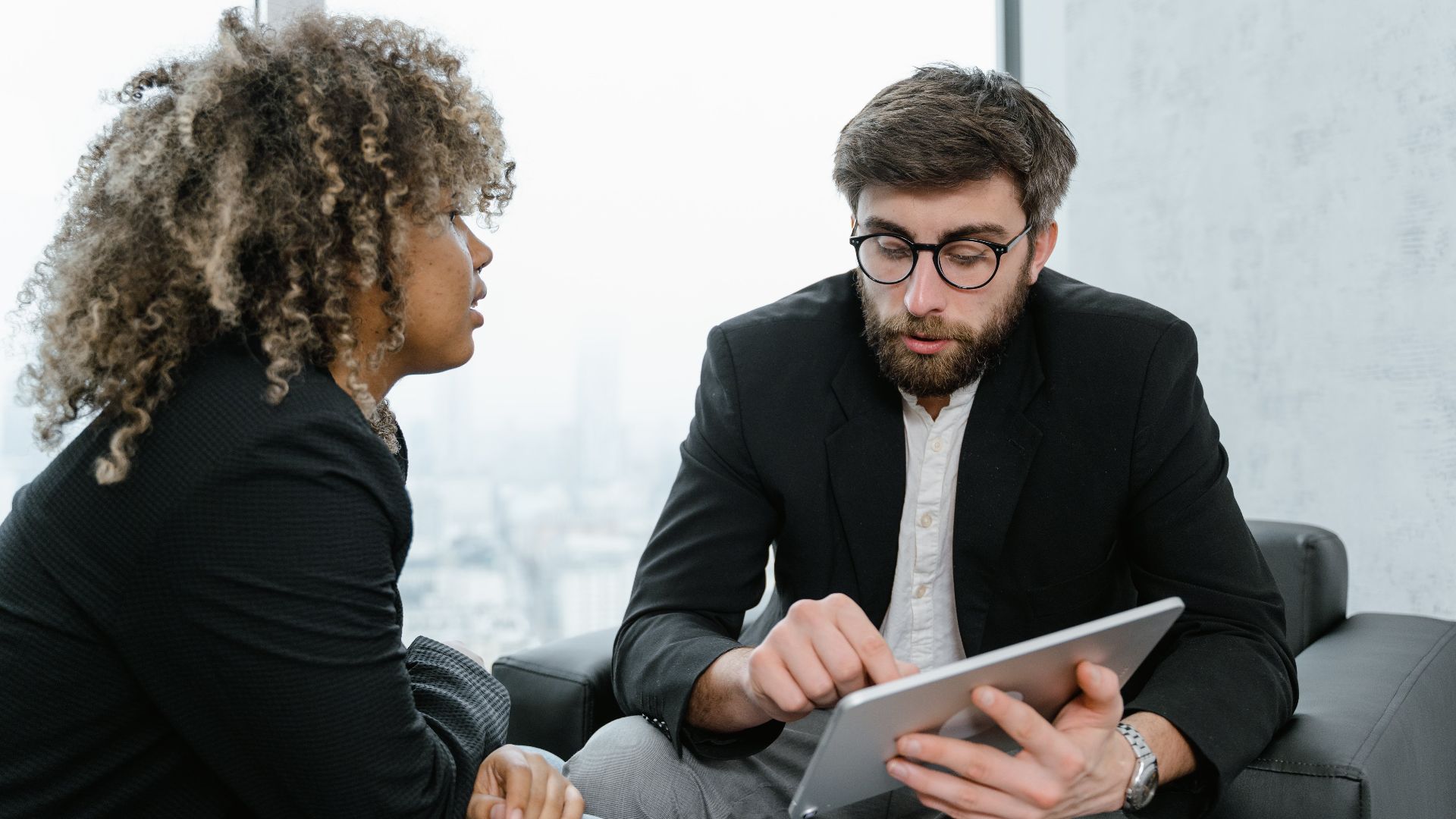 A focused business meeting between two professionals discussing data on a tablet indoors.